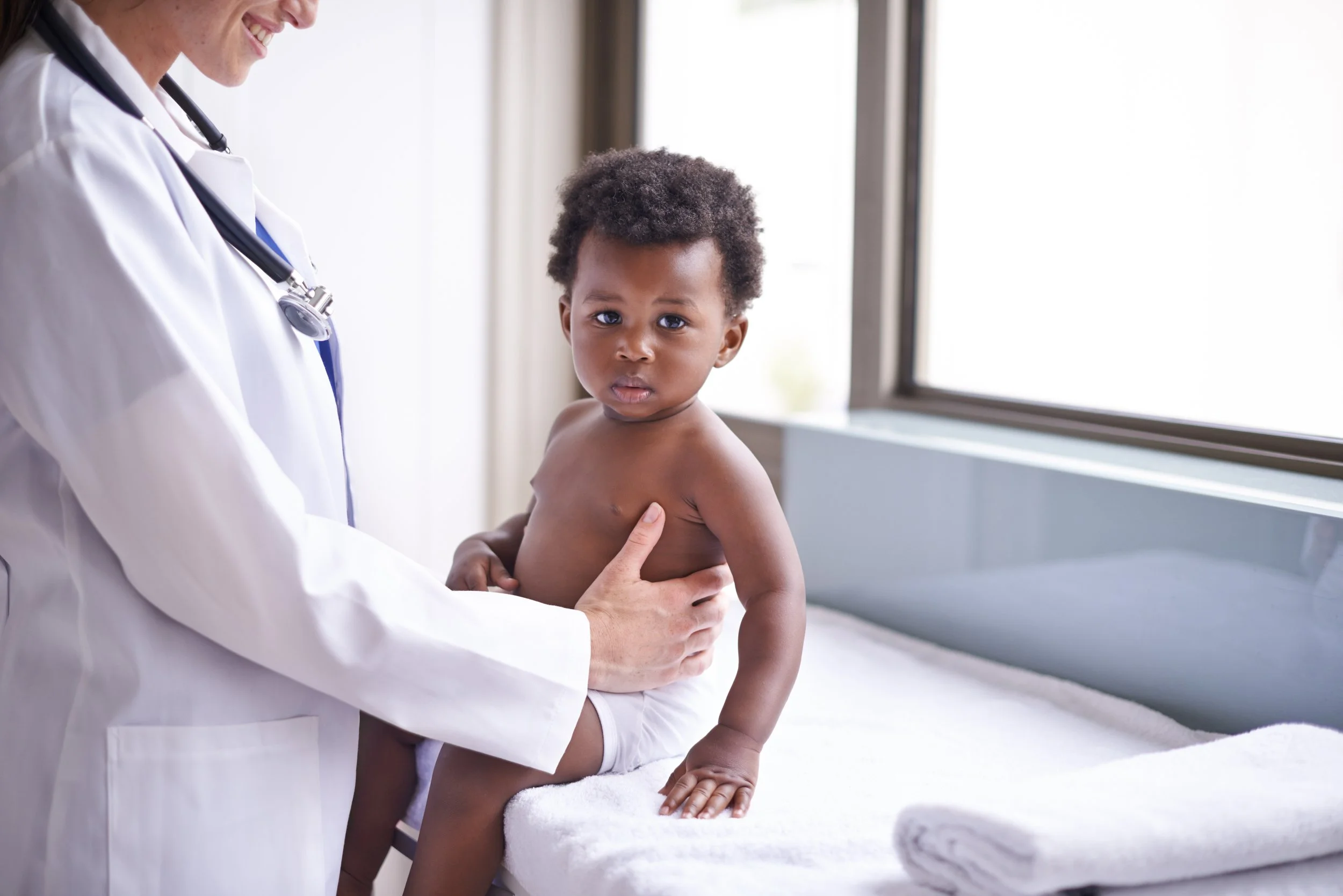 A smiling healthcare professional, wearing a white coat and stethoscope, examining a young boy with curly hair at a medical clinic.