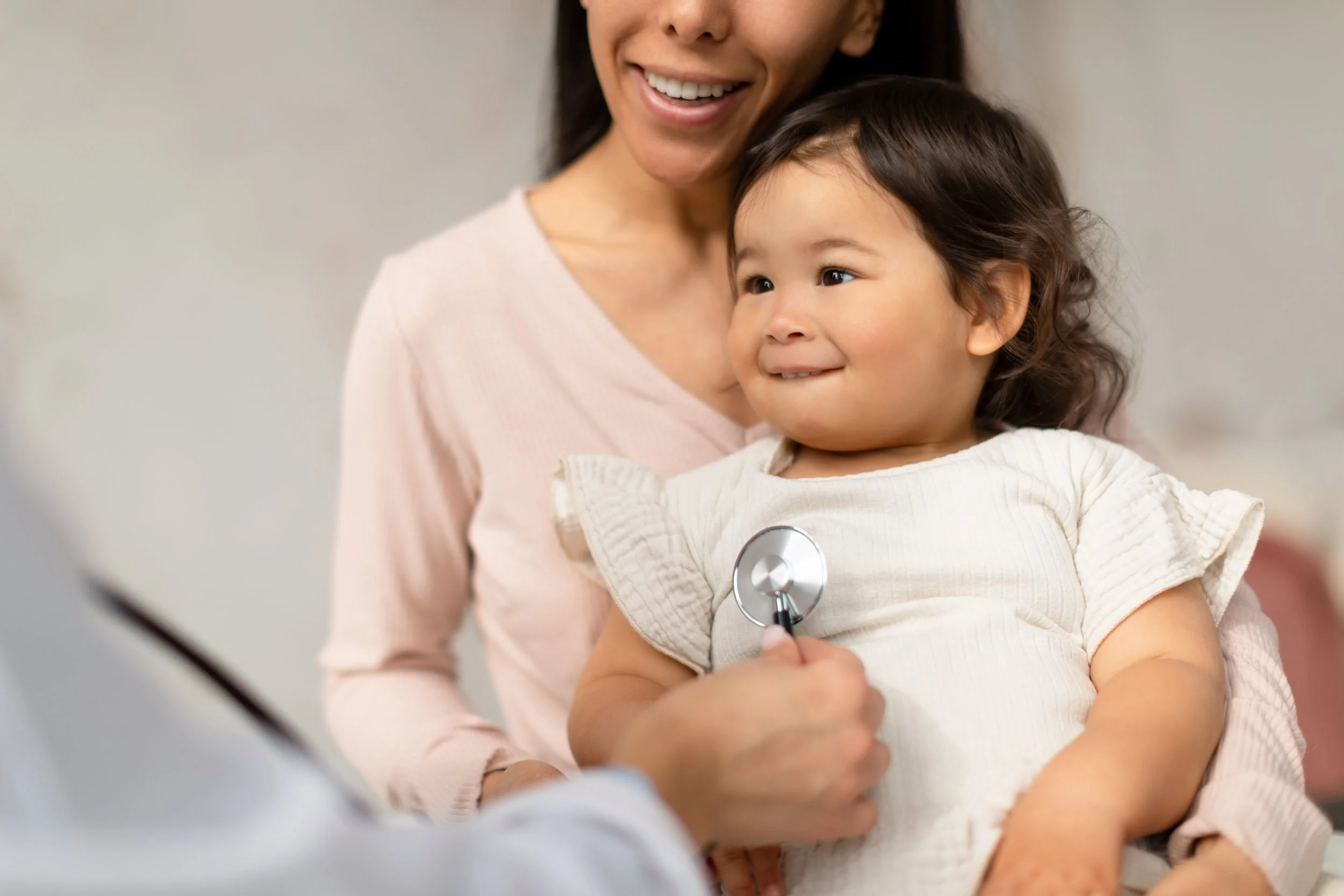 A smiling woman wearing a light pink top is holding a young girl in a white top with ruffled sleeves; the girl has a stethoscope on her chest and is looking to the side with a slight smile.