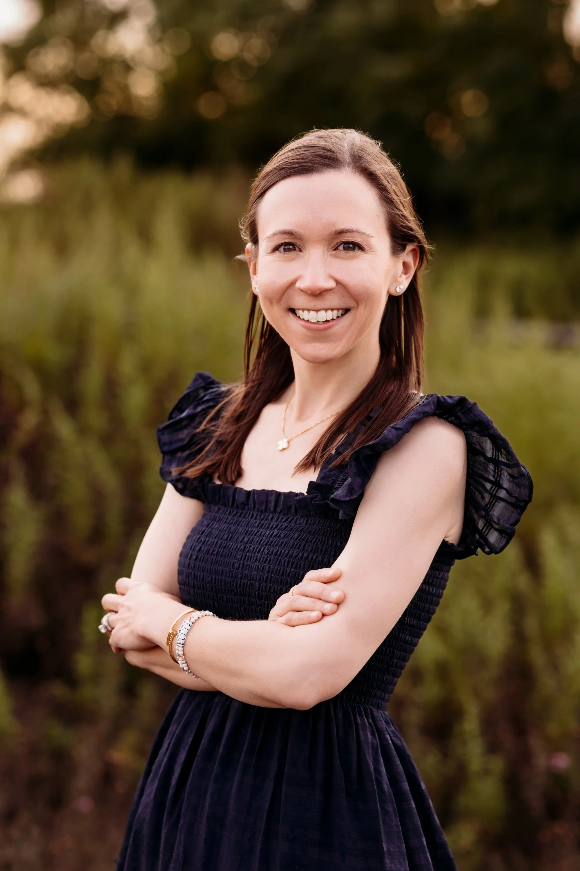 A woman with brown hair and light skin, smiling and standing outdoors with her arms crossed, wearing a navy blue dress with puffed sleeves and various jewelry, in a natural setting with blurred trees and greenery in the background.