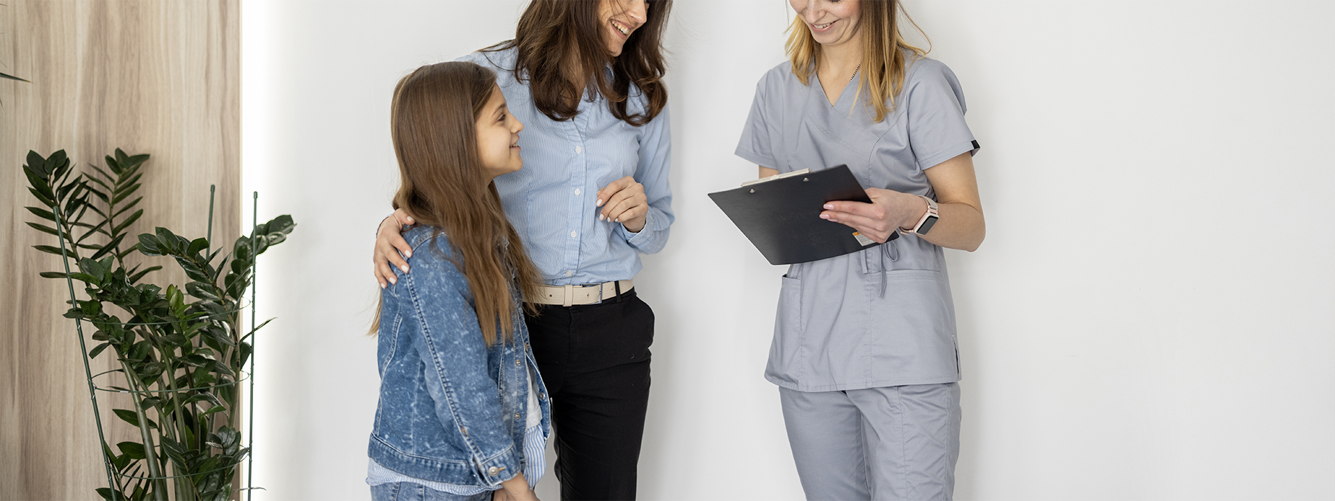 A young girl and two women, one in medical scrubs and one in business casual, smiling during a conversation in a medical or office setting.