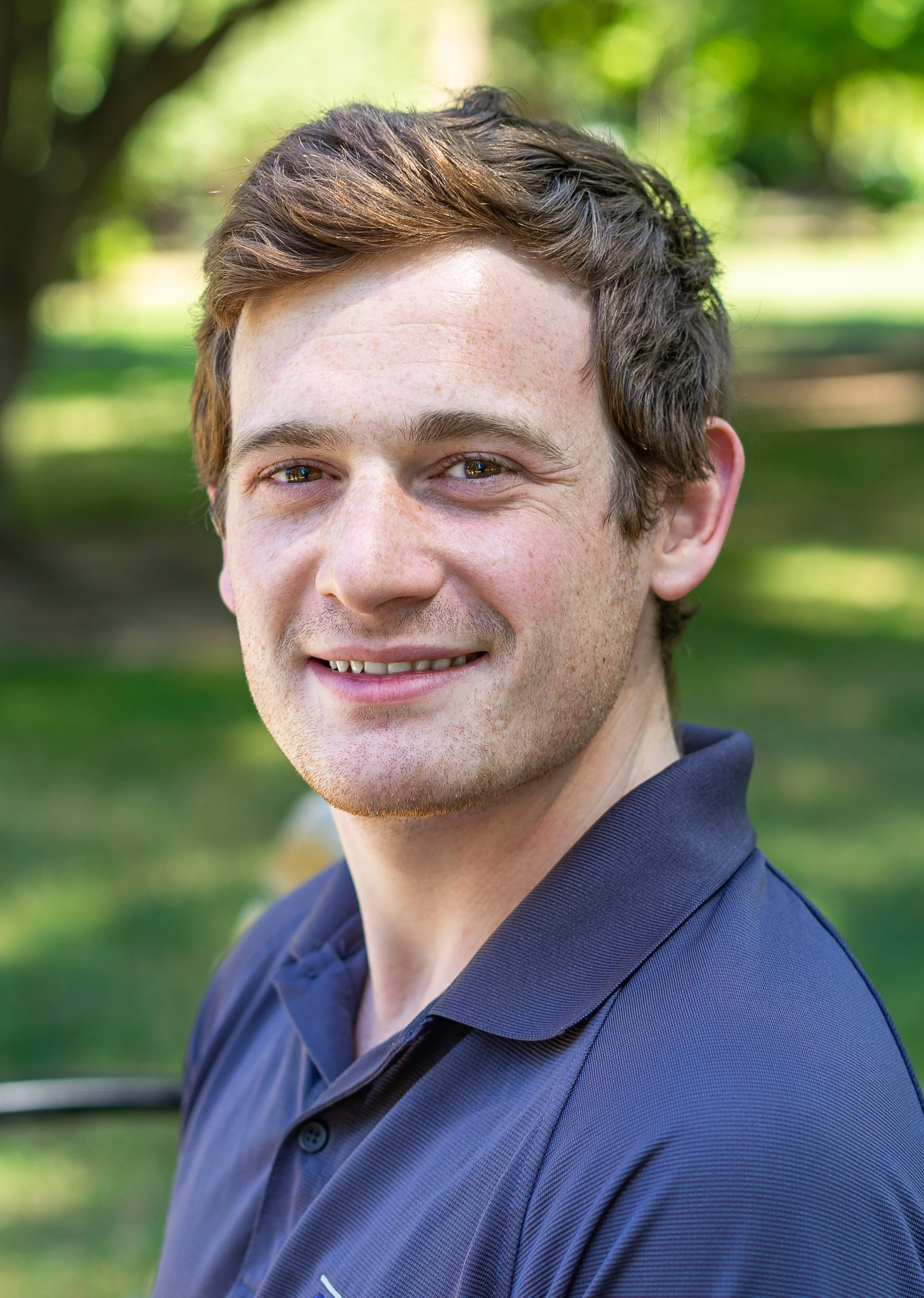 A young man with brown hair, smiling, outdoors in a park with green trees and grass, wearing a dark blue collared shirt.
