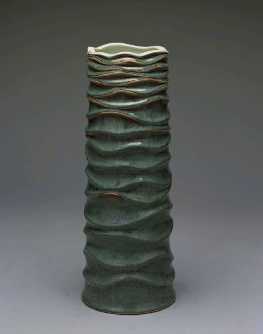 Ceramic sculpture with a wavy, textured surface in shades of green and brown, set against a neutral background.