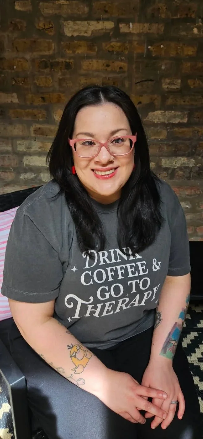 A woman with black hair, pink glasses, and tattoos on her arms, smiling at the camera, sitting in front of a brick wall, wearing a dark gray t-shirt that says 'Drink Coffee & Go to Therapy'.