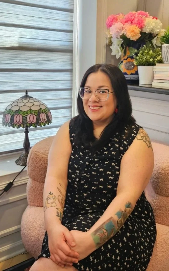 A woman with shoulder-length black hair, glasses, and tattoos on her arms, sitting on a pink plush chair in a cozy room with a large window, a Tiffany-style lamp, a bouquet of pink, white, and peach flowers, and a stack of books in the background.