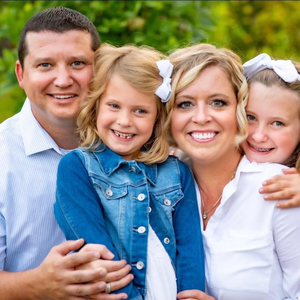 A smiling family of four, including a man, a woman, and two young girls, outdoors with green trees in the background.
