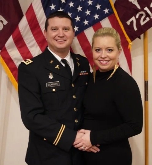A man in military uniform and a woman in black dress standing together, holding hands, with American flags in the background.