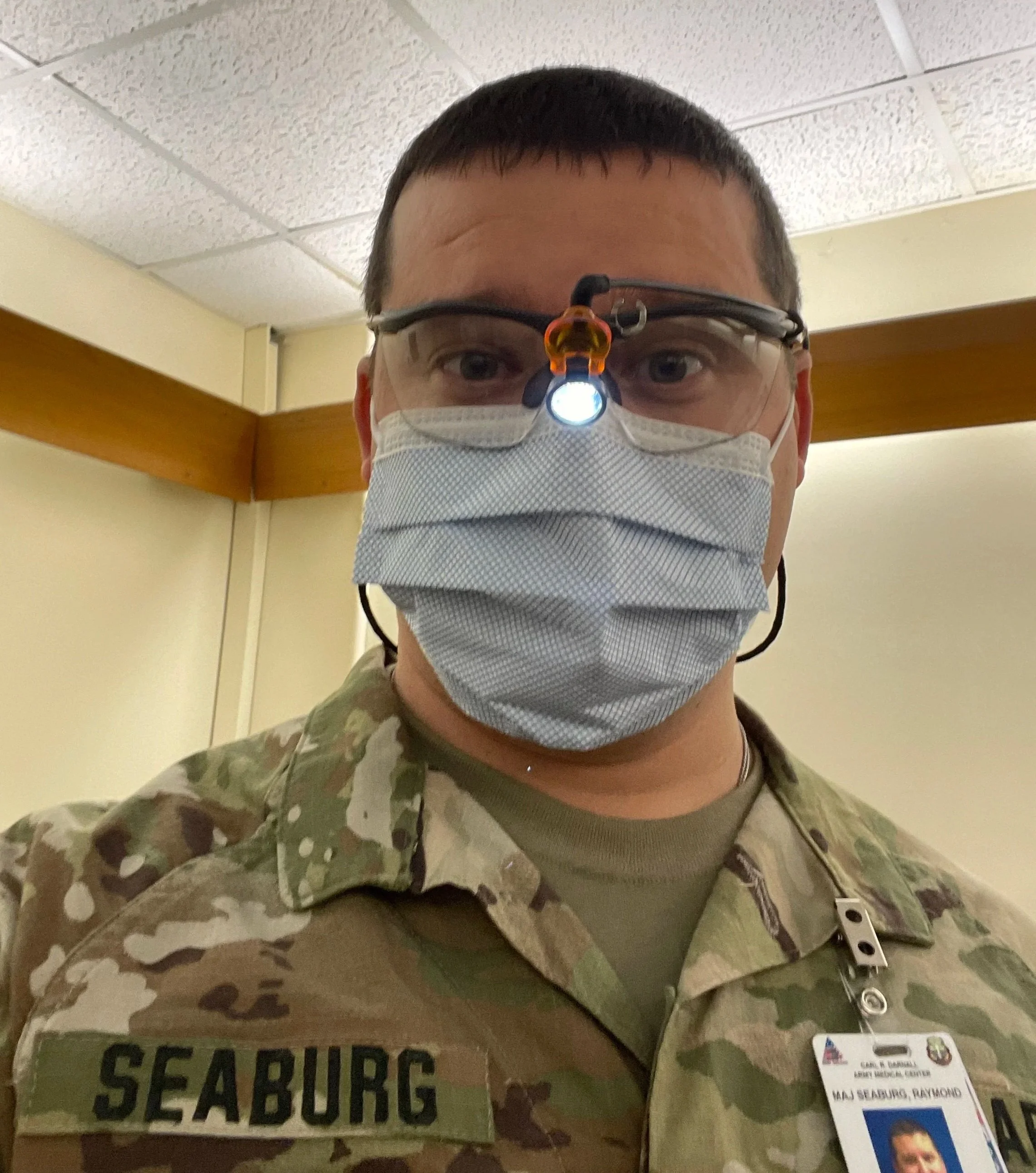 A person wearing military camouflage uniform, safety glasses with a headlamp, and a face mask, standing indoors with a SAFBUEG sign in the foreground and a badge with a photo, name, and rank.