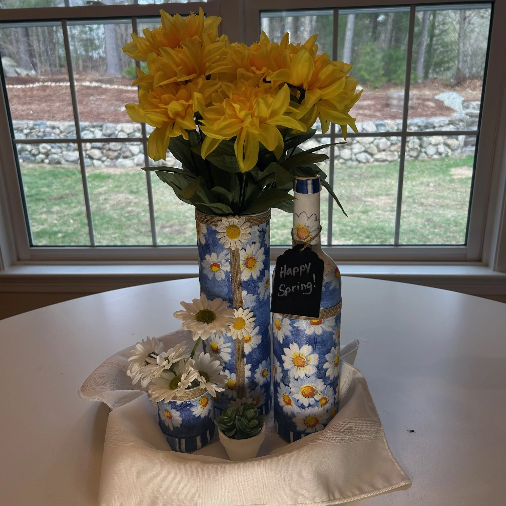 A bouquet of yellow flowers, including daisies, in a decorated vase with a sunflower pattern, placed on a table in front of a window overlooking a backyard.