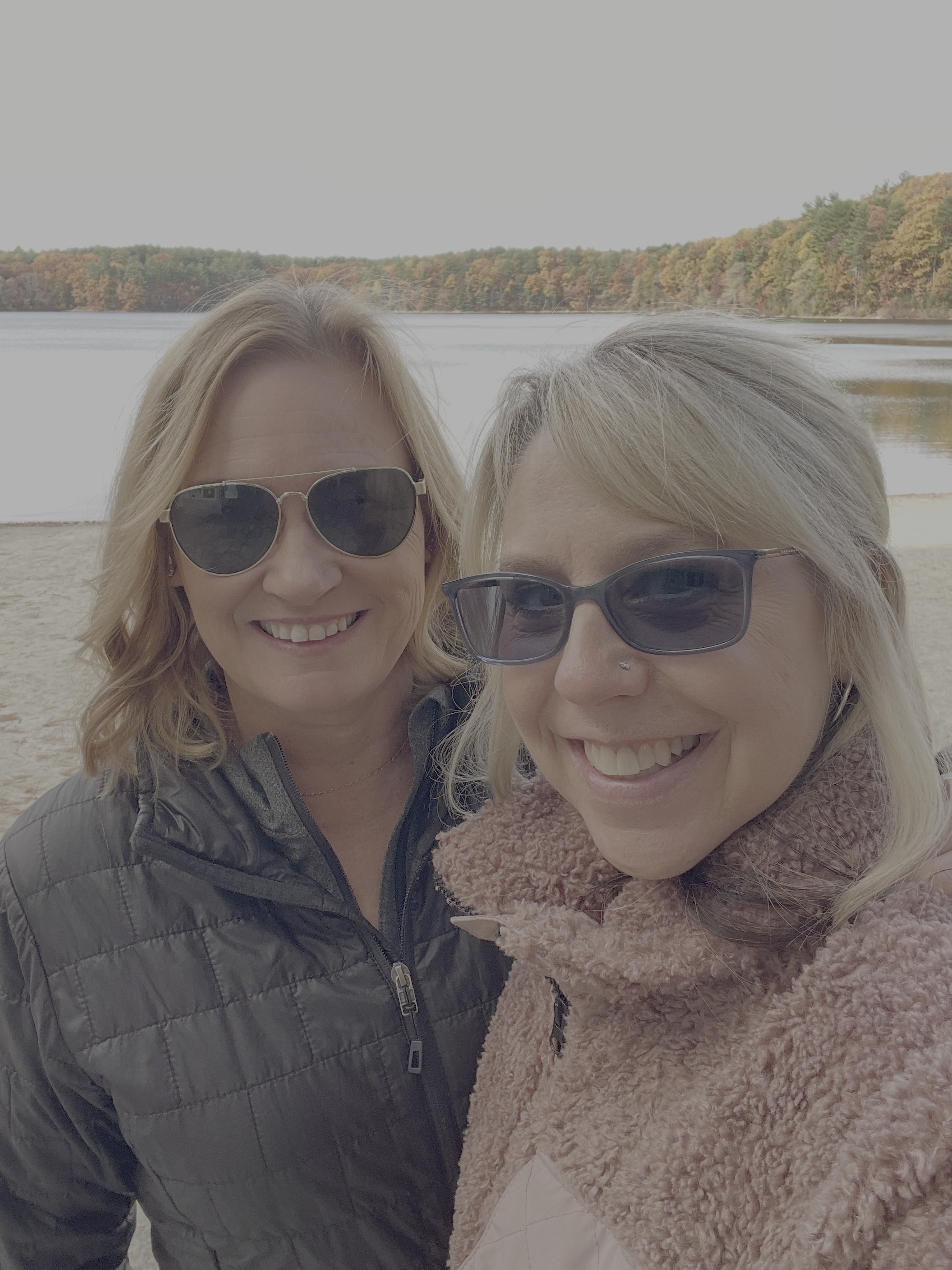 Two women smiling outdoors by a lake with a forested shoreline in autumn, both wearing sunglasses and jackets.