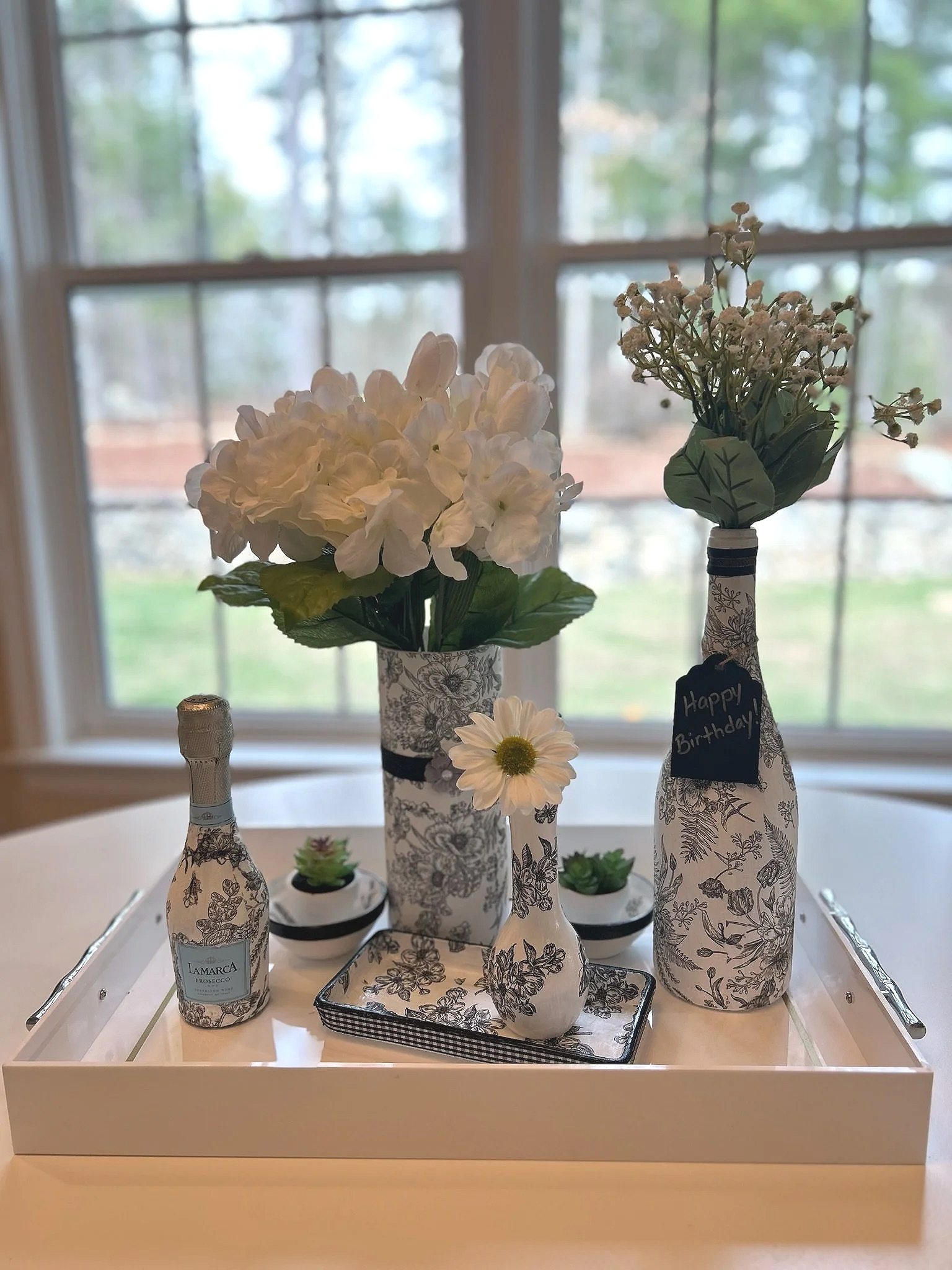 A birthday display on a tray with decorative bottles wrapped in floral-patterned paper, containing flowers and small plants, with a window showing greenery in the background.