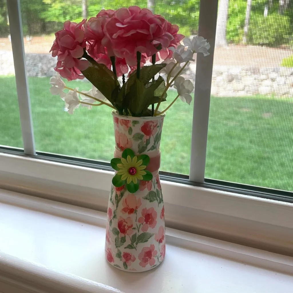 A floral patterned vase holding pink and white artificial flowers on a windowsill with a screen, overlooking a green yard.