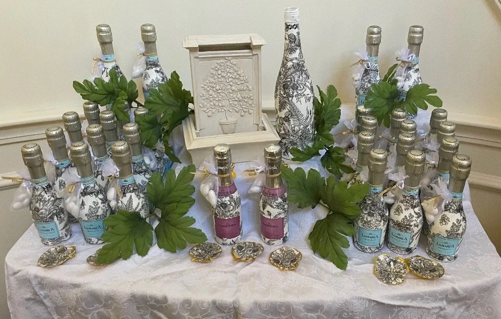 Decorative display of bottles wrapped with floral patterned paper, set on a white tablecloth with green leaves; includes a small white fountain and a ceramic bottle with a black and white botanical design, surrounded by seashells.