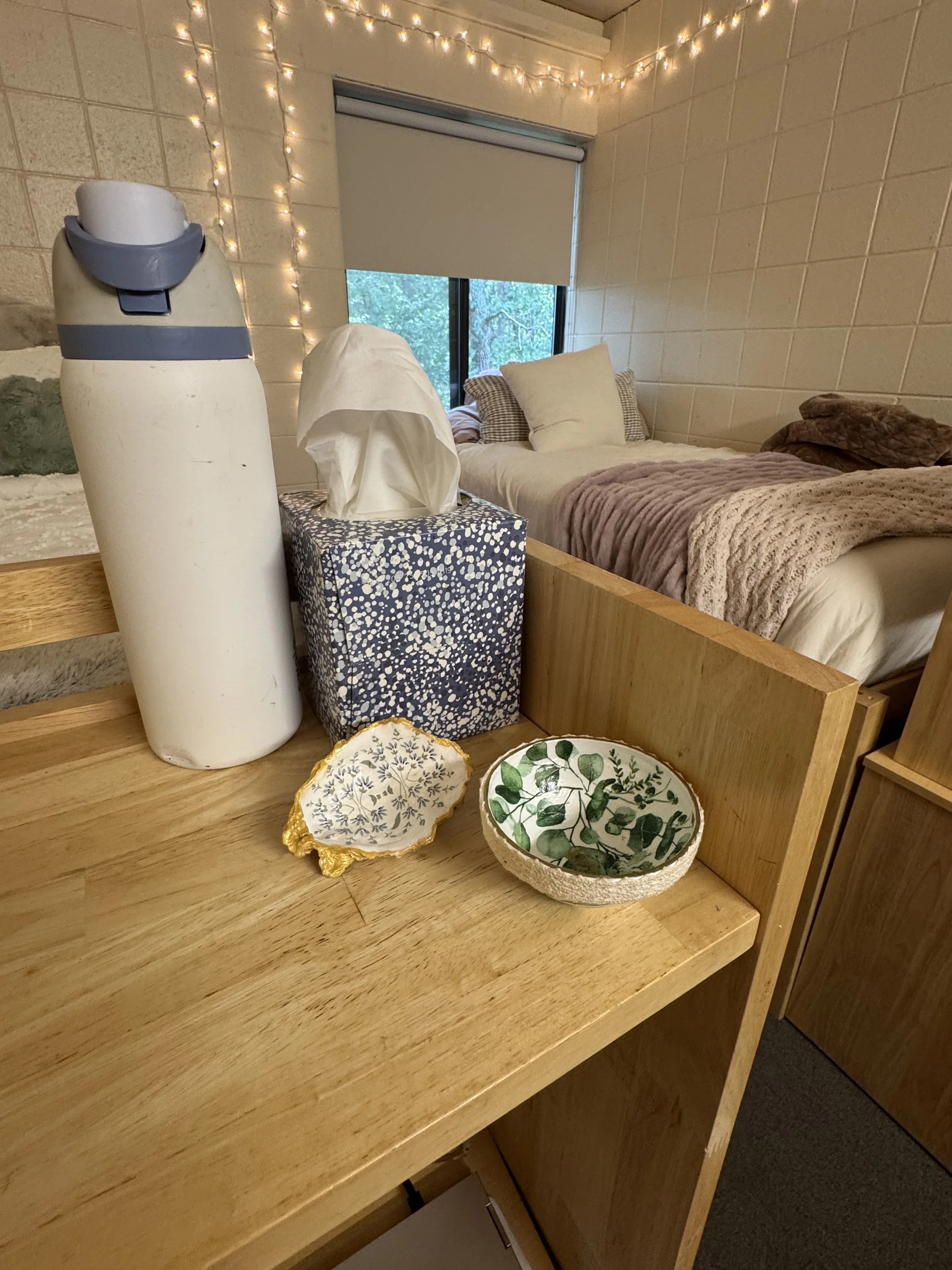 A corner of a room with a bed, fairy lights, a window with a rolled-up blind, a tissue box, and decorative bowls on a wooden surface.
