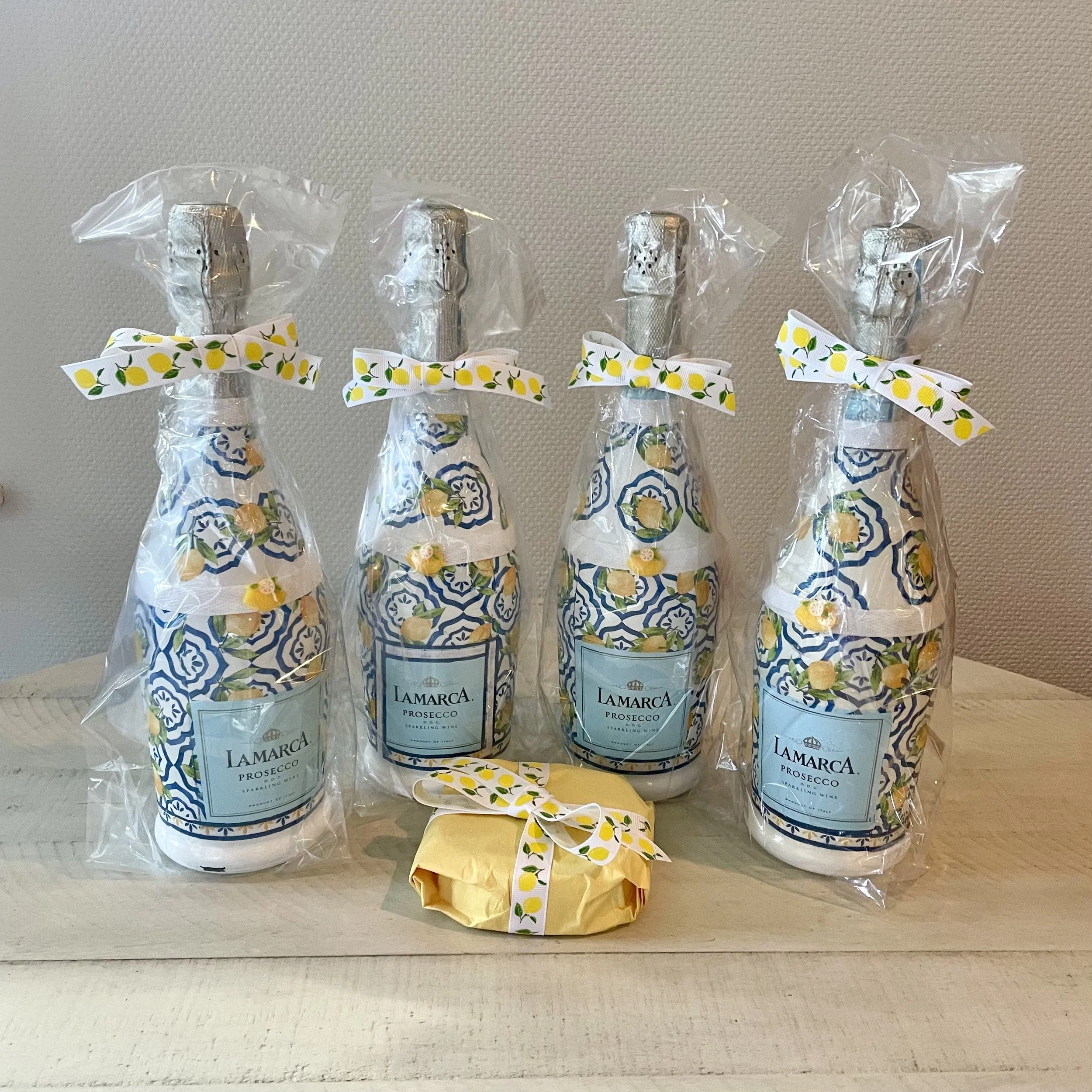Four bottles of Lamarca Prosecco sparkling wine, wrapped in clear plastic with decorative lemon-themed ribbons, standing on a light-colored surface against a beige textured wall.