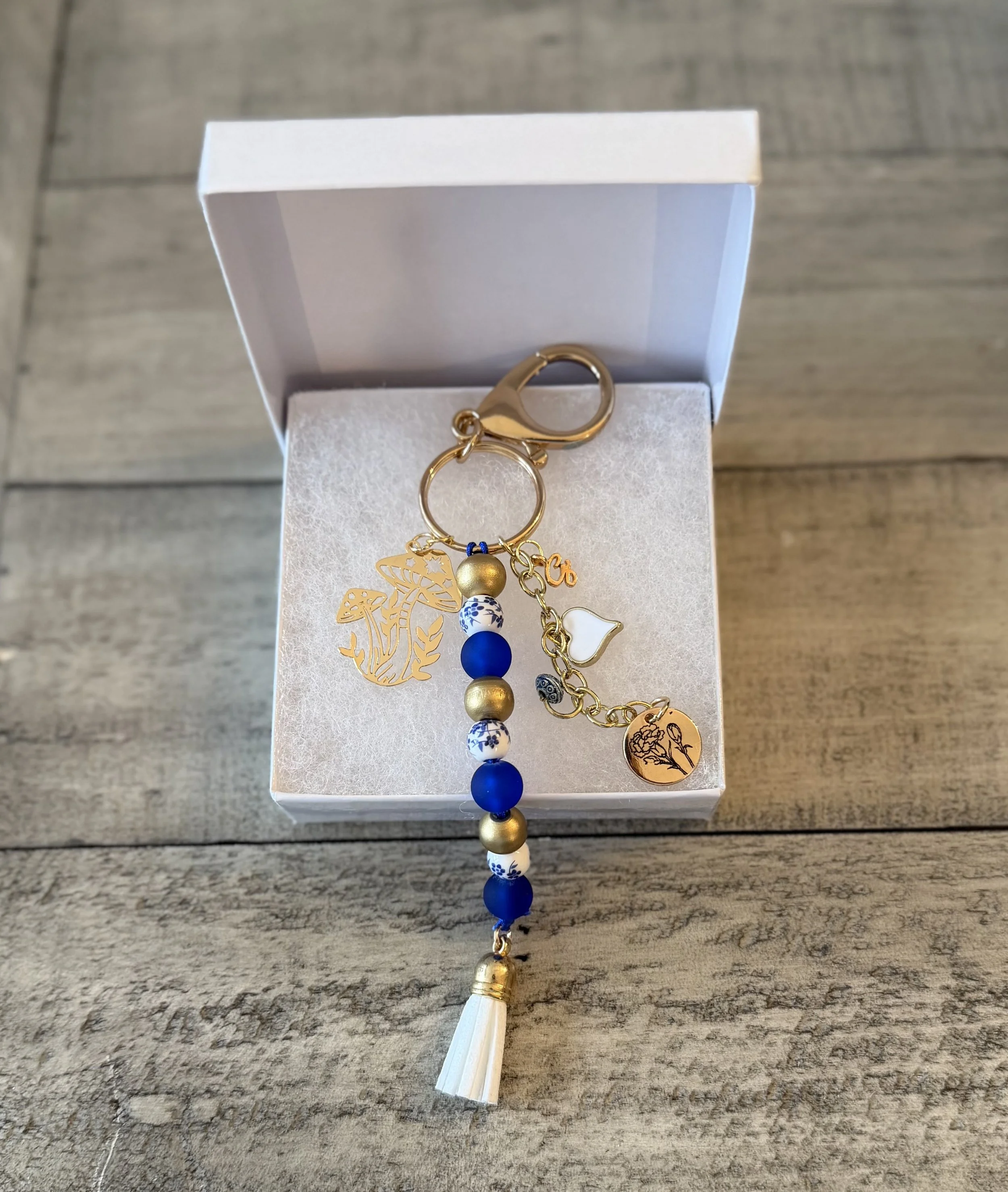 A white jewelry box with a gold charm keychain inside, placed on a wooden surface. The keychain has various charms including gold, white, and blue beads, a butterfly leaf design, a bird charm, and a disc with a floral design. The keychain has a white tassel attached at the bottom.