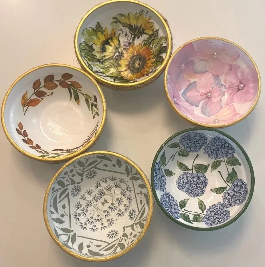 Five decorative bowls with floral and leaf patterns, each with a gold rim, arranged on a light-colored surface.