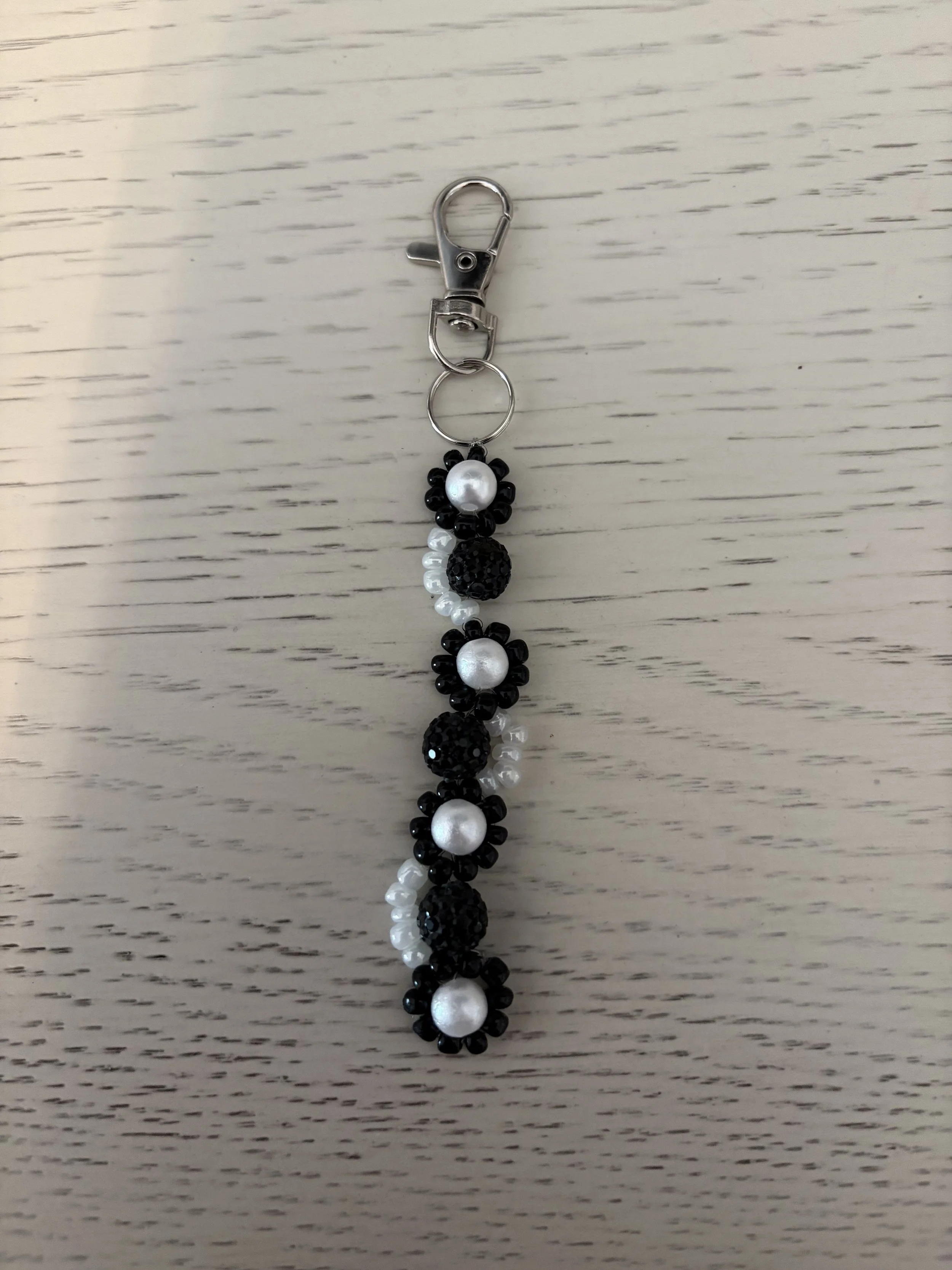 A beaded keychain with black and white beads, including large white pearls and smaller black beads, hanging from a metal clip on a white wooden surface.