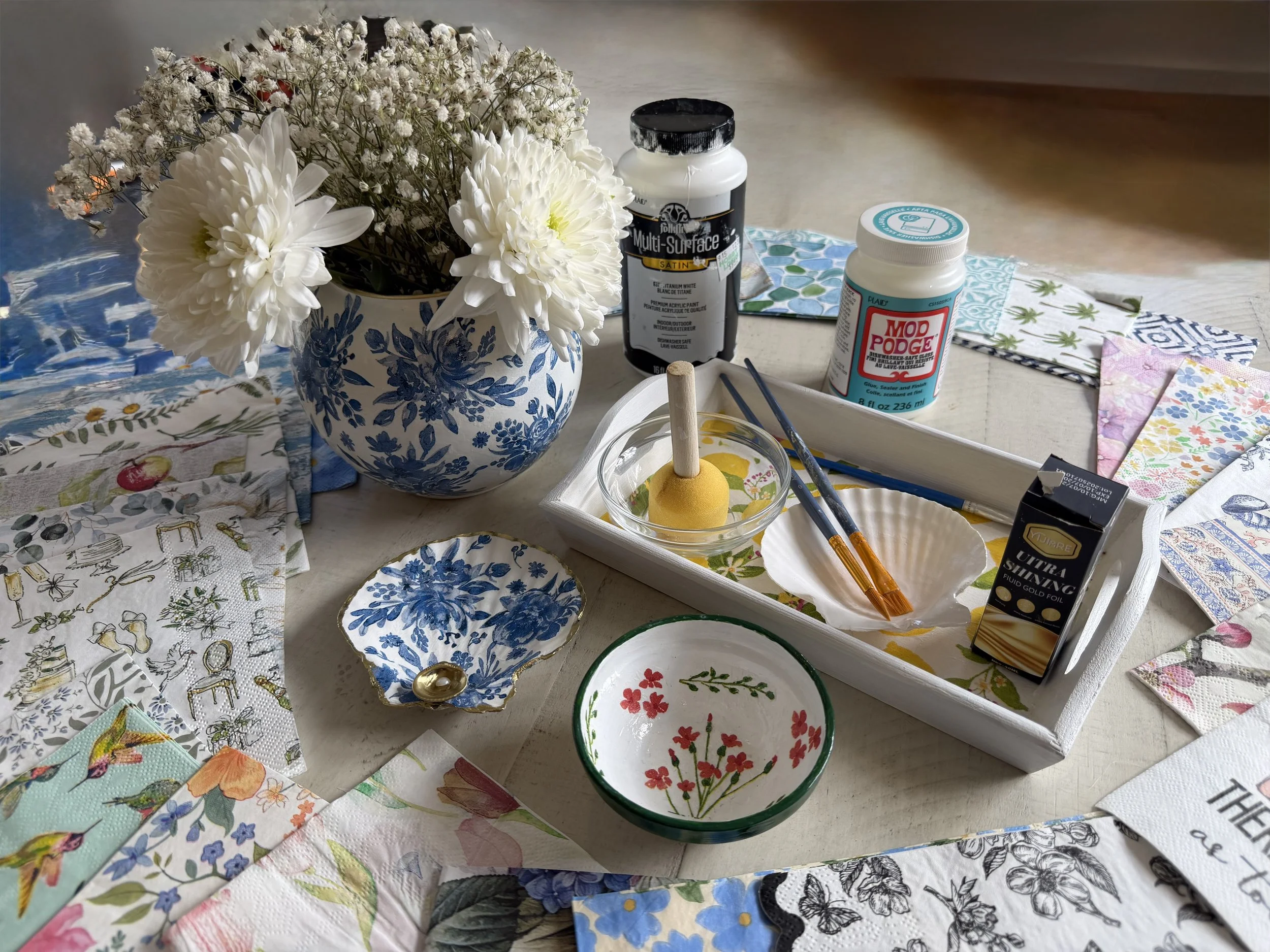 Assorted arts and crafts supplies on a table, including a bouquet of white flowers in a blue and white vase, paper napkins with various patterns, small decorated bowls, acrylic paint bottles, paint brushes, and foil paper.