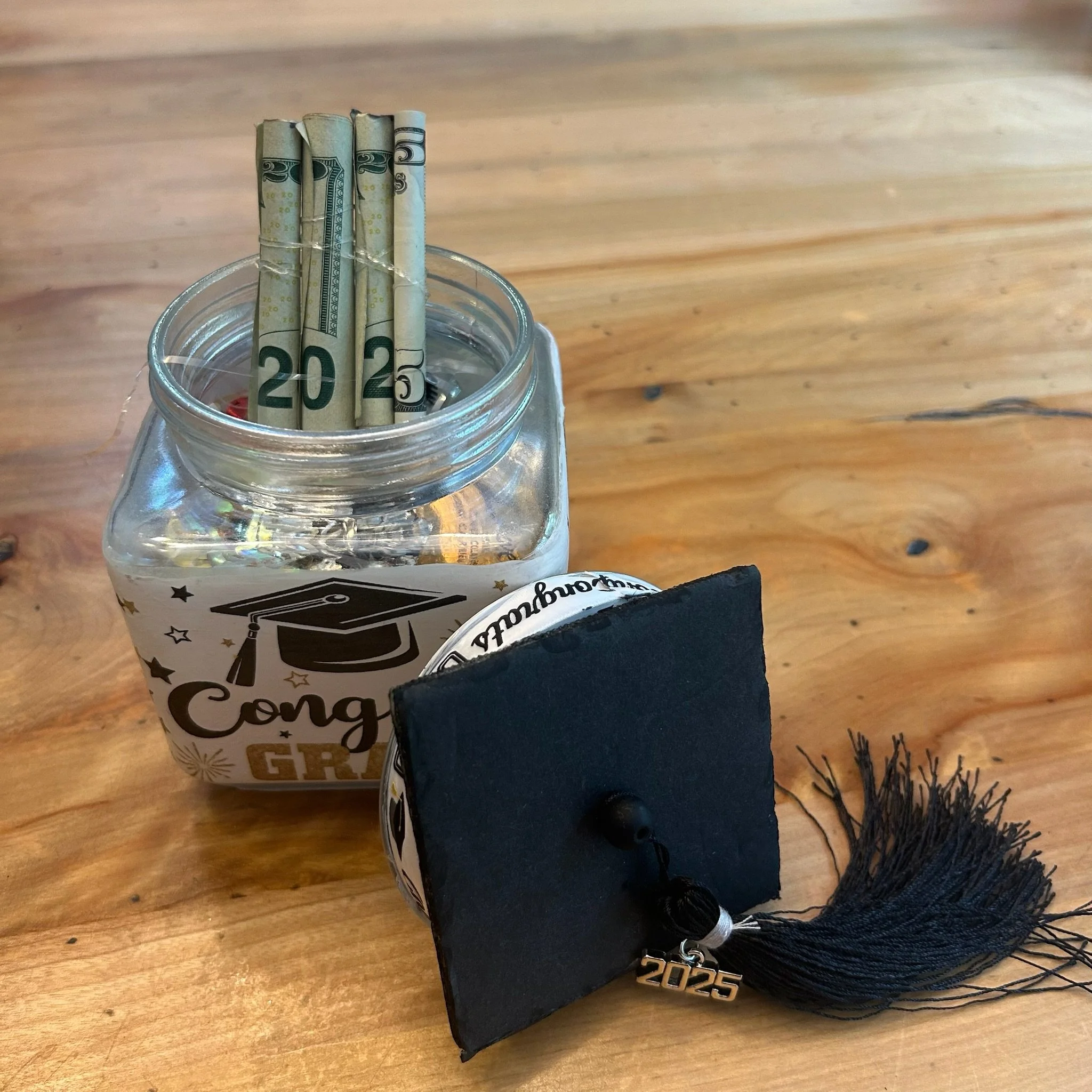 A glass jar with rolled twenty dollar bills attached with rubber bands, sitting on a wooden table. In the foreground, a black object with a '2025' keychain is visible, and in the background, part of a black item with fringes and handwritten text can 