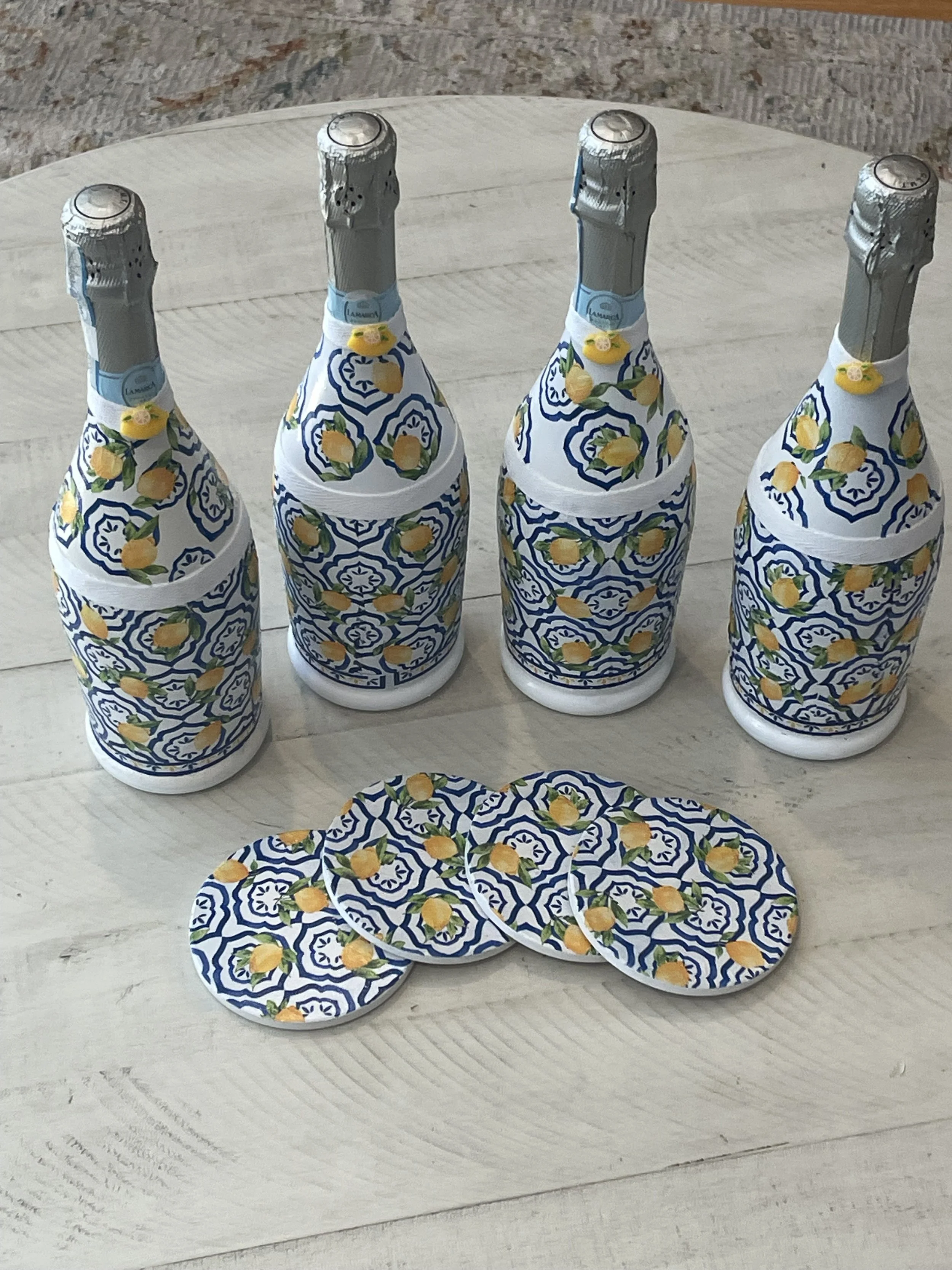 Four bottles of sparkling water wrapped in decorative paper with yellow lemons and blue patterns, placed on a light-colored wooden surface with three matching coasters.
