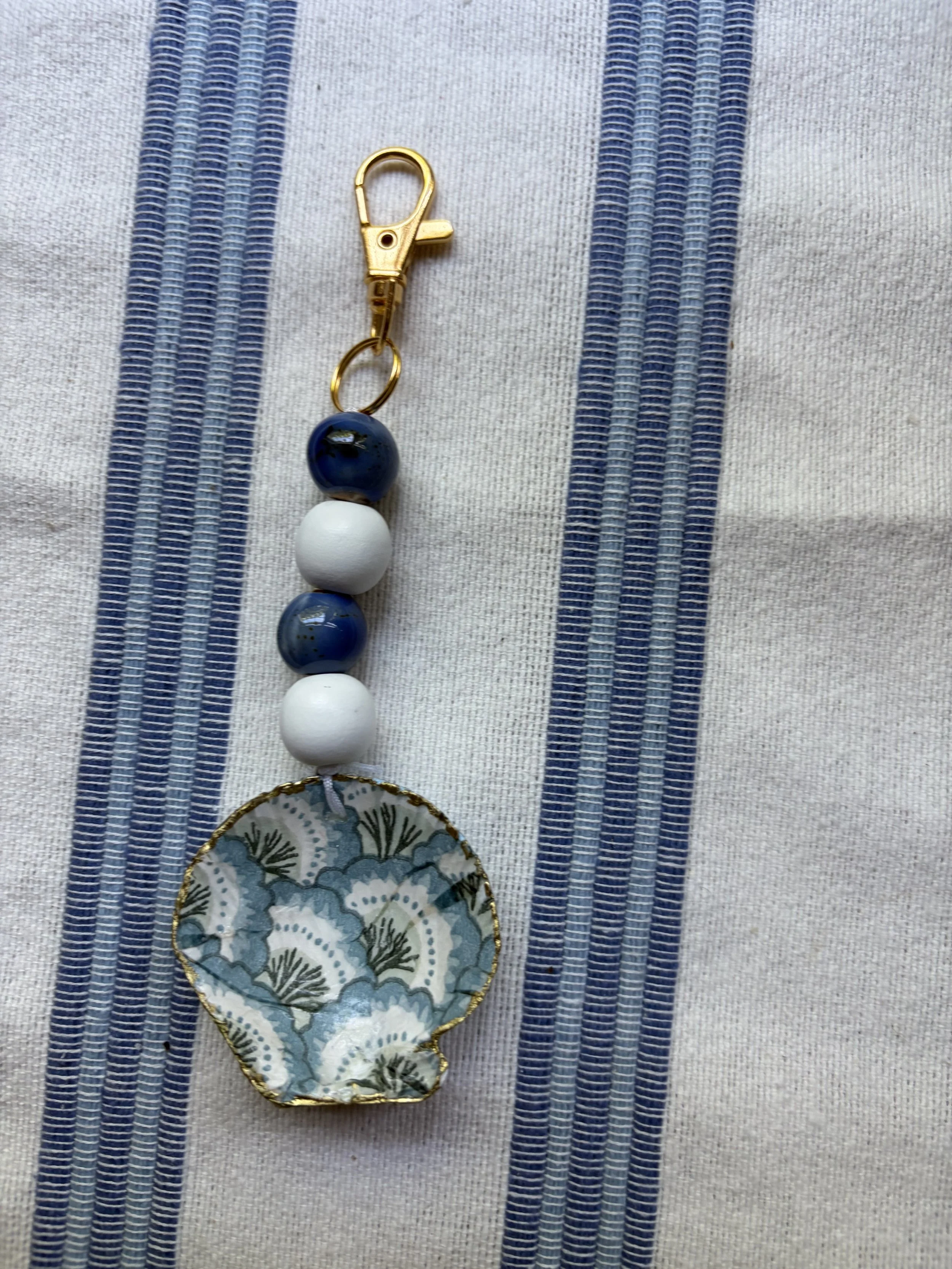 A decorative keychain with a floral and botanical design, featuring white and blue beads and a fabric pendant, placed on a cloth with blue and white stripes.