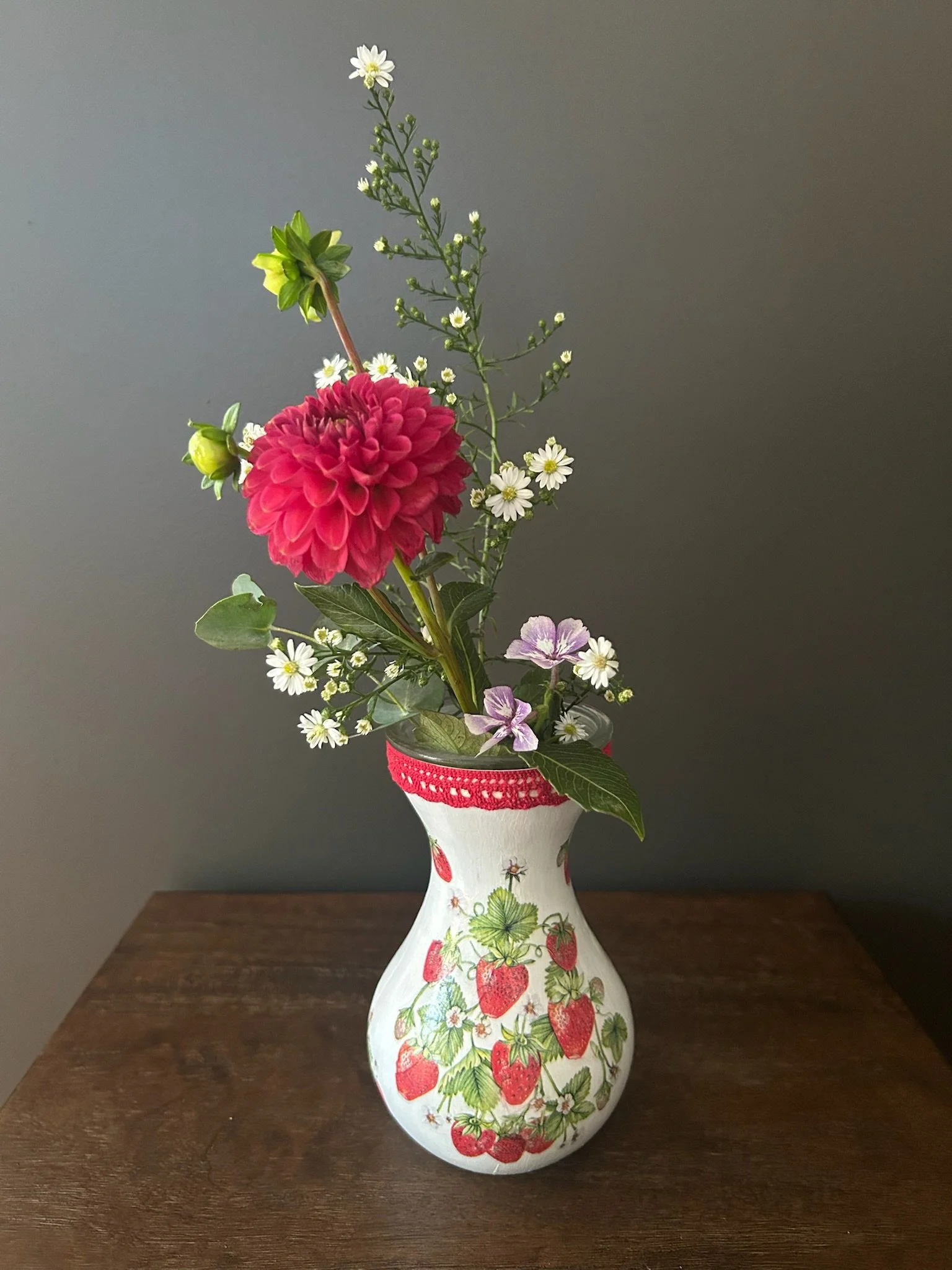 A bouquet of various flowers in a strawberry-themed vase on a wooden surface against a plain background.