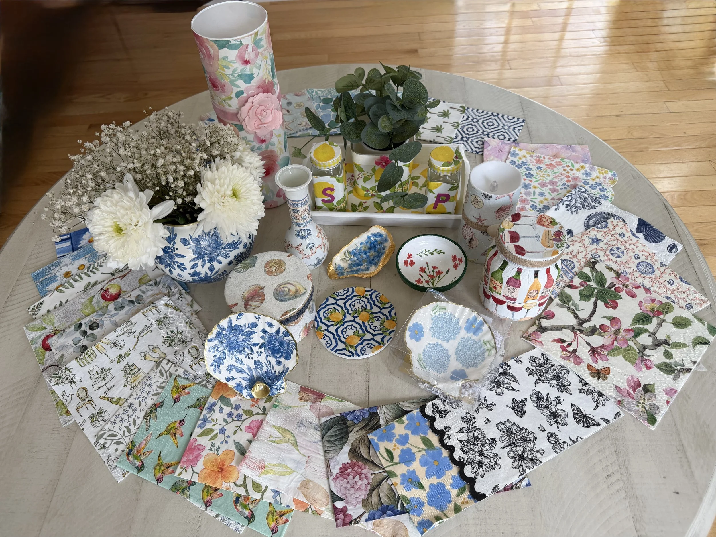 A collection of floral-patterned paper napkins, a floral vase with white flowers, a green leafy plant in a white container, smaller decorated jars, and decorative plates on a light wooden table with a hardwood floor background.