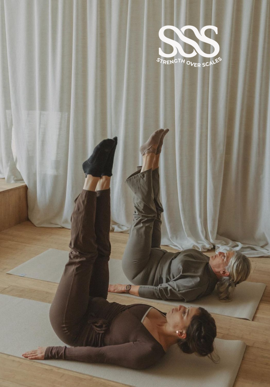 Two women practicing yoga on mats inside a room with beige curtains. They are lying on their backs with their legs raised and feet pointing upward. The logo "Strength Over Scales" is in the upper right corner.