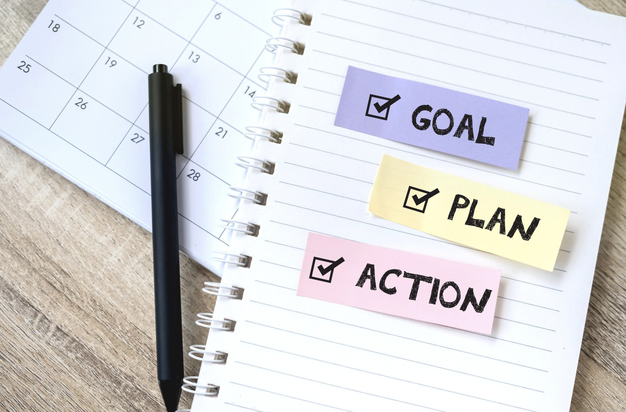 Een agenda met gekleurde plakbriefjes met de woorden 'GOAL', 'PLAN', en 'ACTIVITY', een zwarte pen, en een spiraalband op een houten tafel.