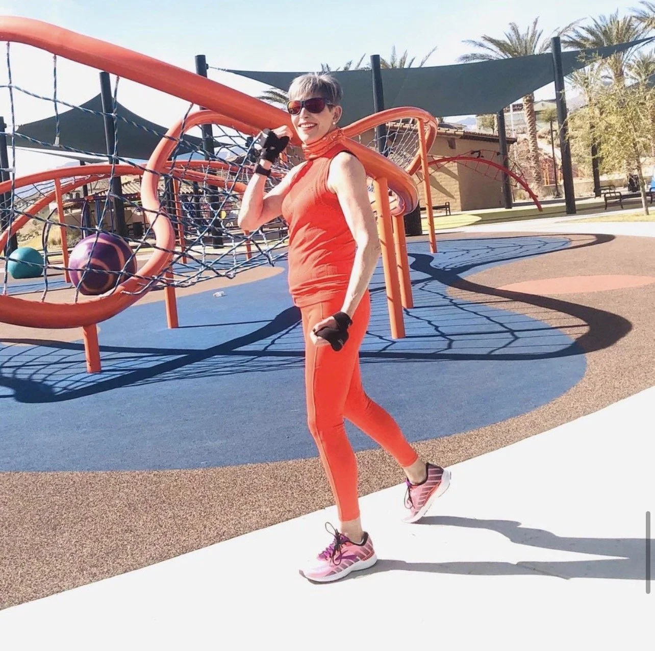 Woman in orange athletic wear and sunglasses at a playground, holding a jump rope, with play structures in the background