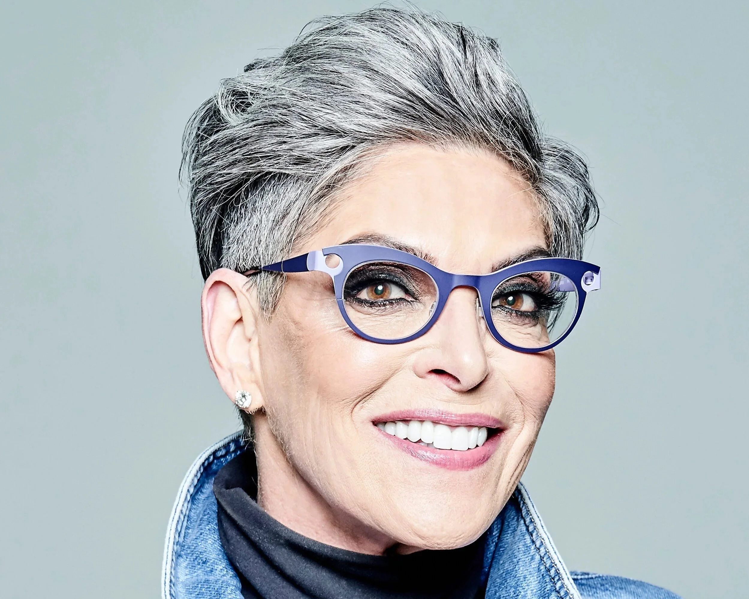 Close-up of a smiling woman with short gray hair, wearing purple glasses, earrings, and a denim jacket.