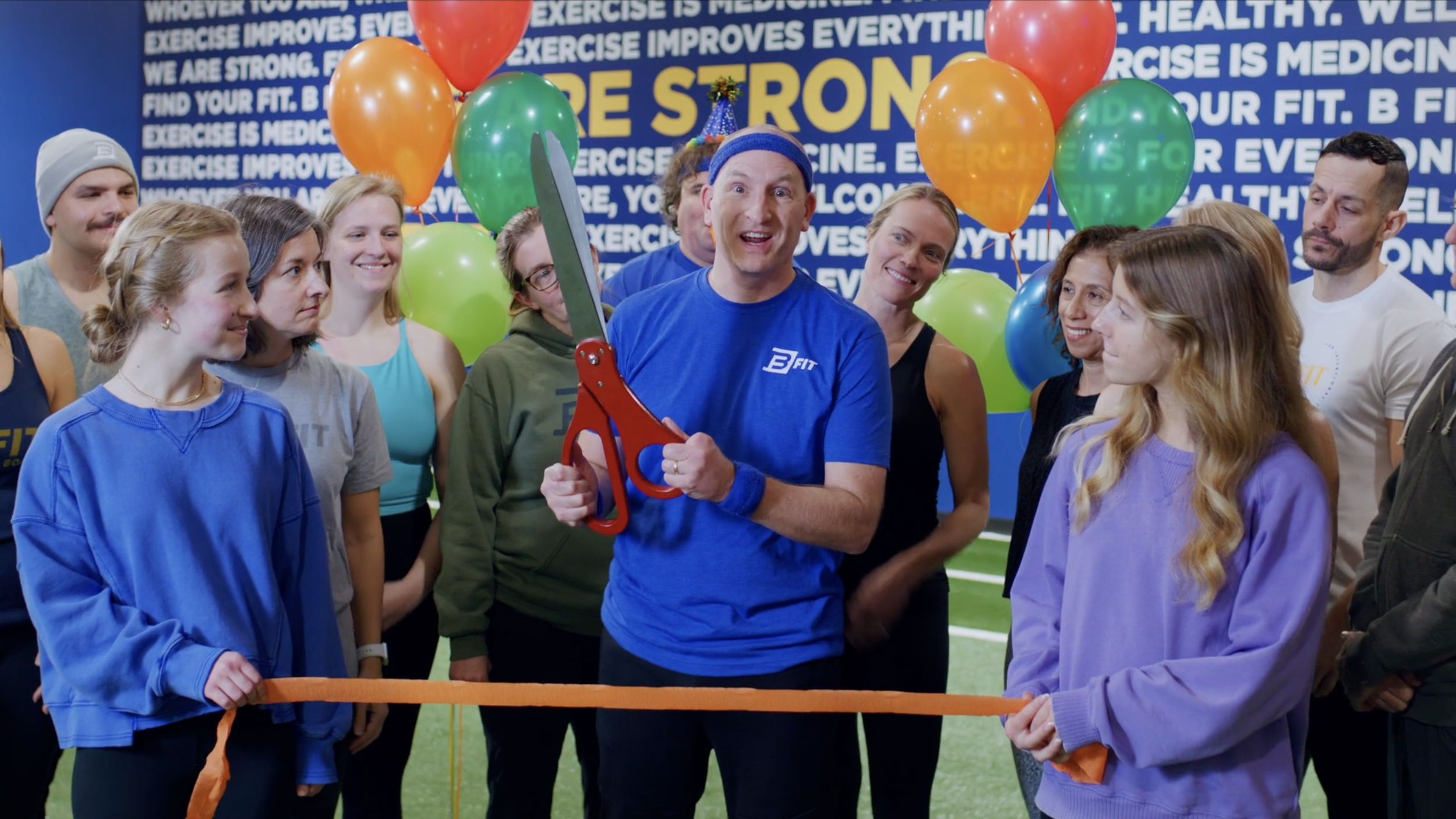 Group of people at a gym or fitness center during a ribbon-cutting ceremony, with colorful balloons and a man holding large scissors.