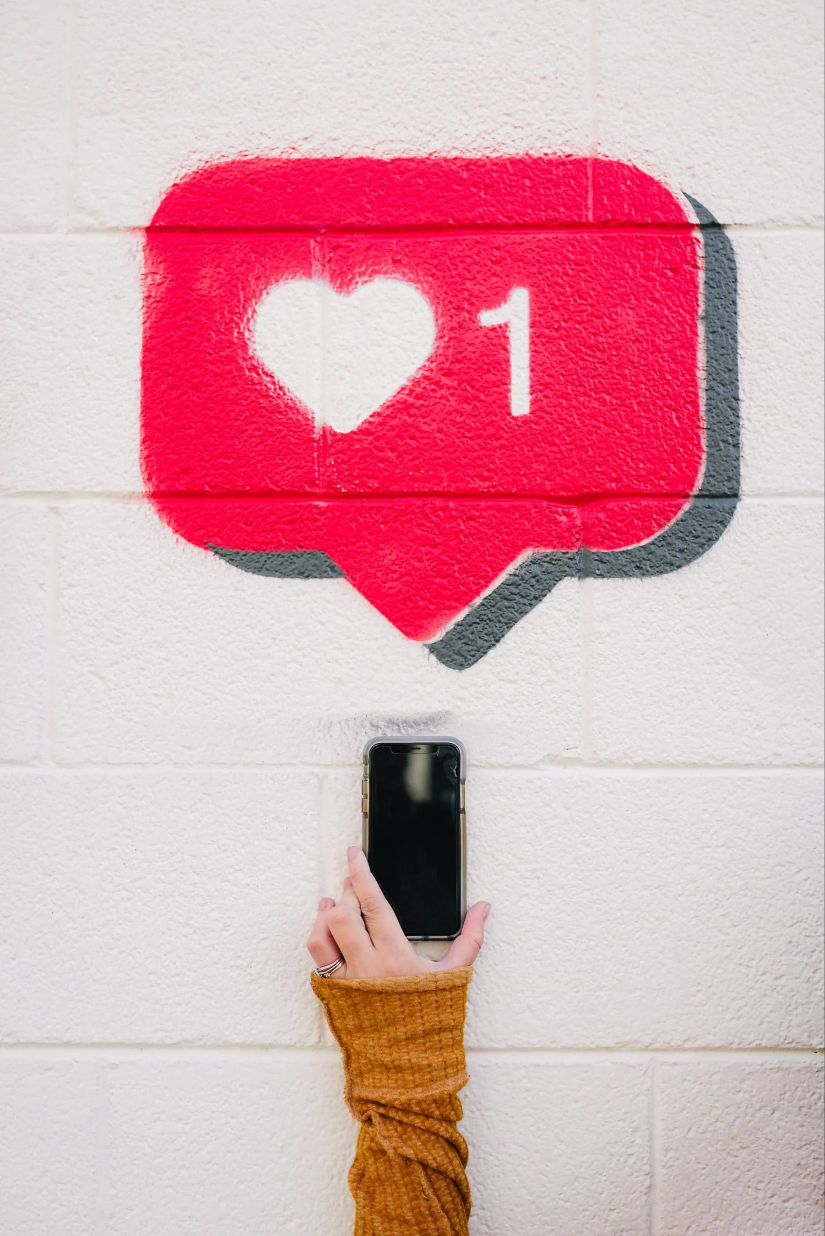 A person with a yellow sweater is holding a smartphone up to a wall art piece that depicts a social media notification icon with a heart and the number 1. The person is standing in front of a brick wall.
