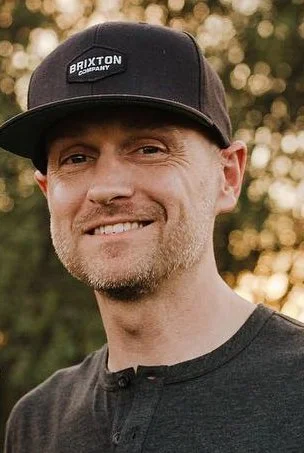 A man wearing a black Brixton cap and a dark gray shirt, smiling outdoors with blurred trees and sunlight in the background.
