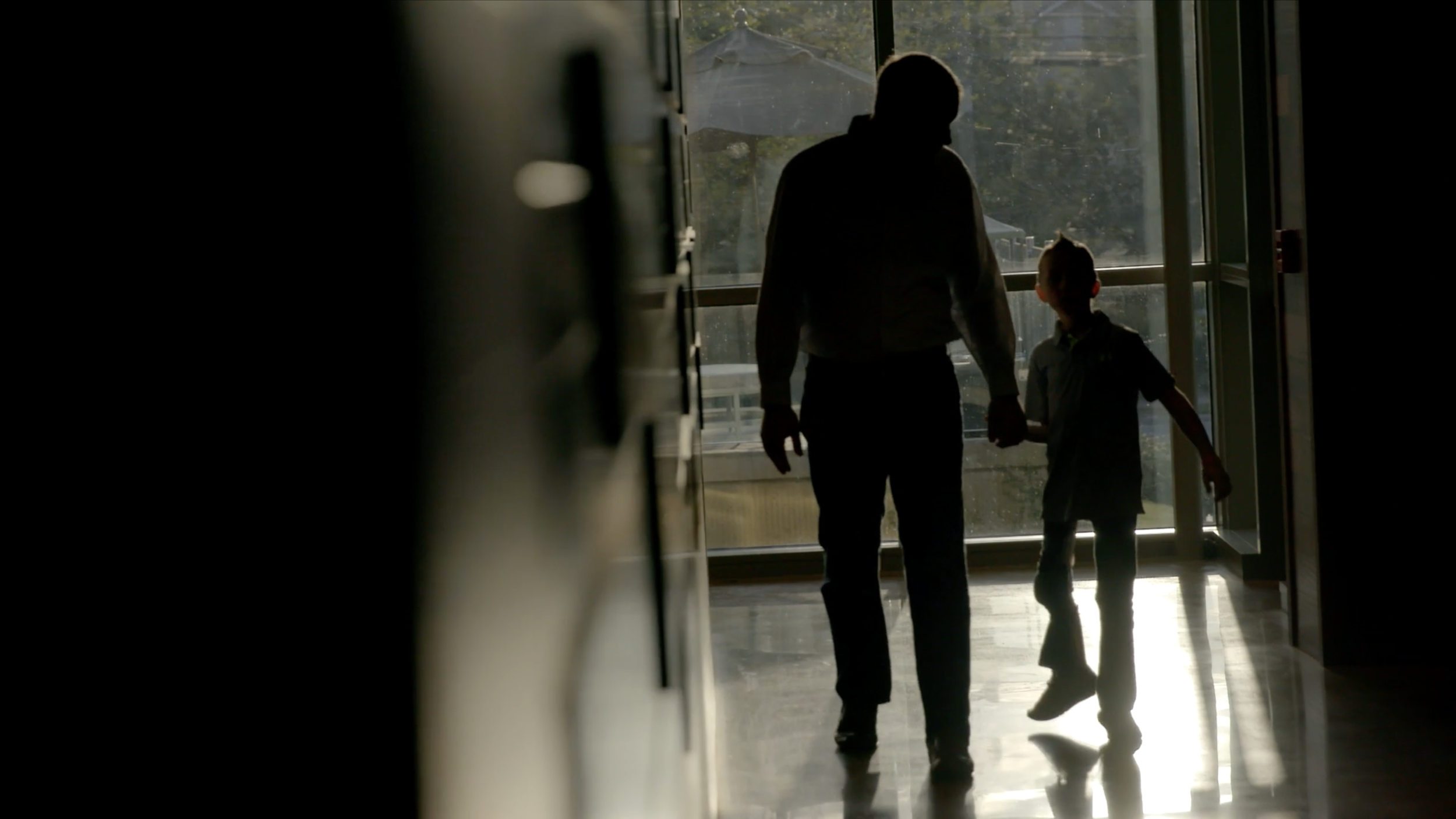 Video production scene of silhouettes of an adult and a child holding hands, walking through a doorway to the outdoors, with sunlight and trees visible outside.