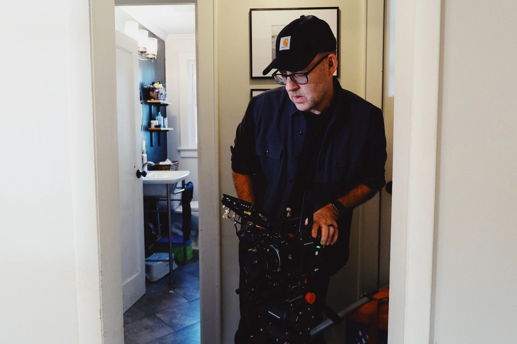 Chris Weatherly operating a camera or filming equipment in a home interior setting, with a bathroom visible in the background.