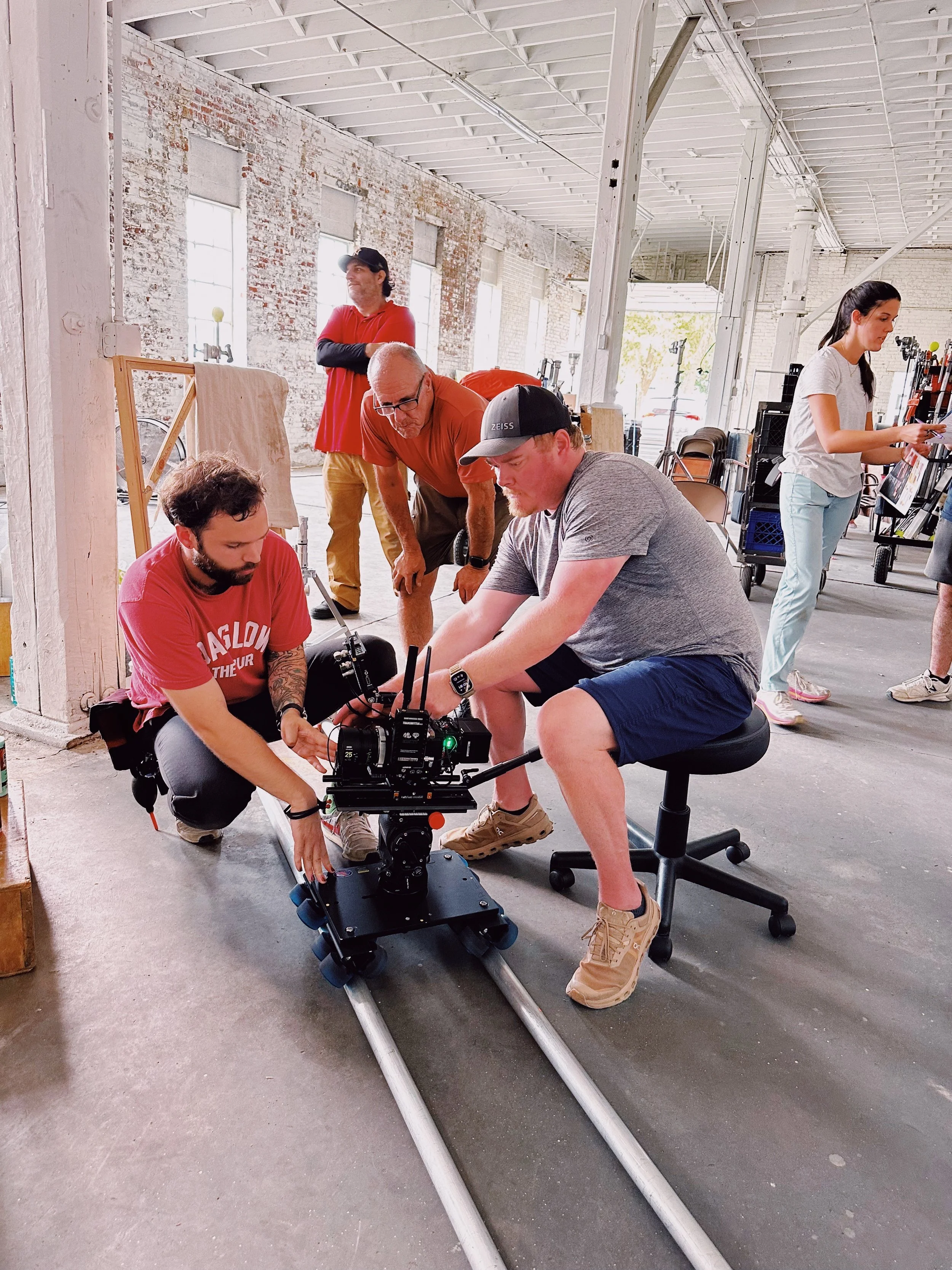 Team of people working together to operate a video camera on a dolly track in a spacious industrial-style studio with brick walls and large windows.