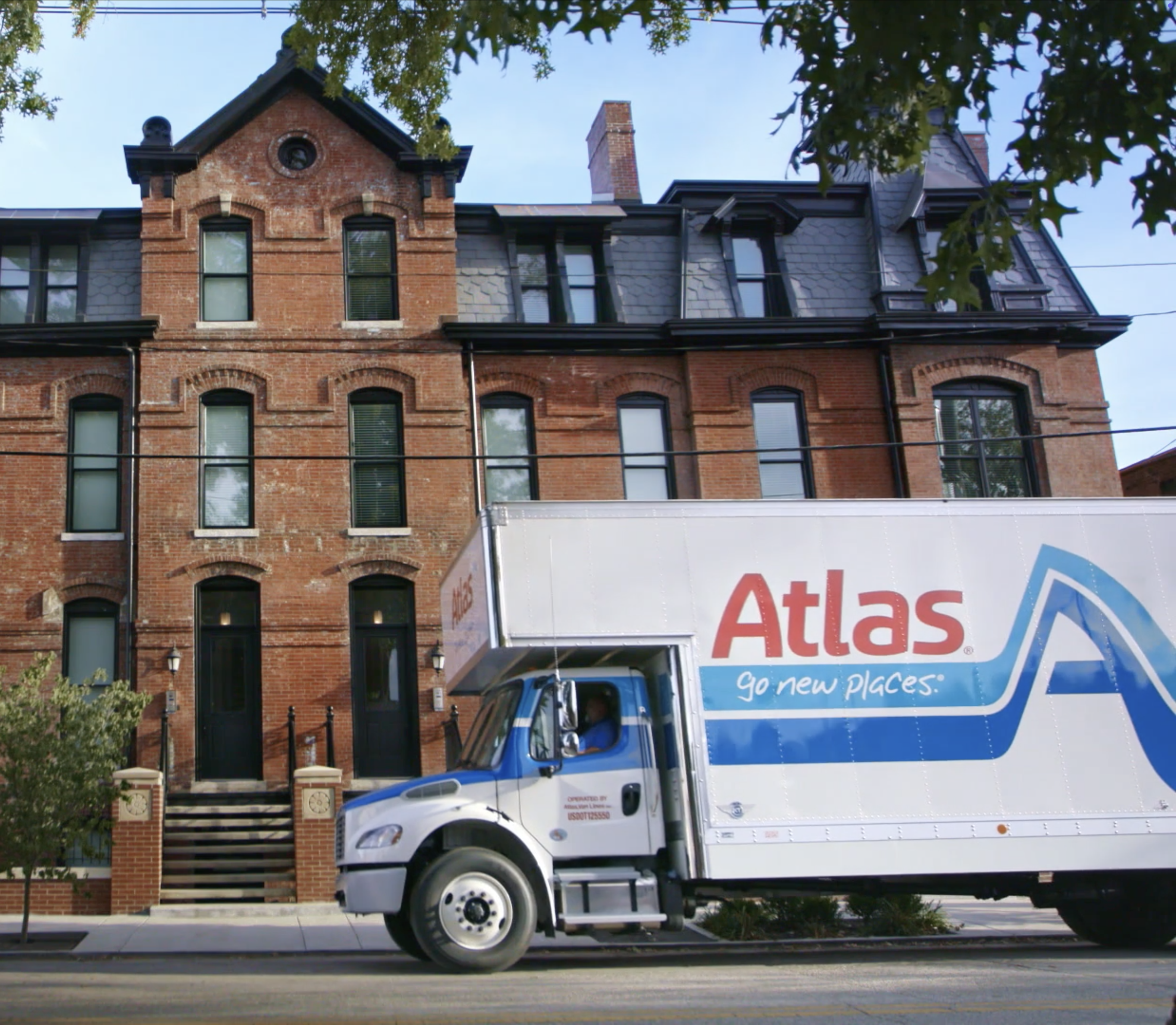 A brick building with multiple windows and dormers on a blue sky background. A blue Atlas moving truck is parked in front of the building.