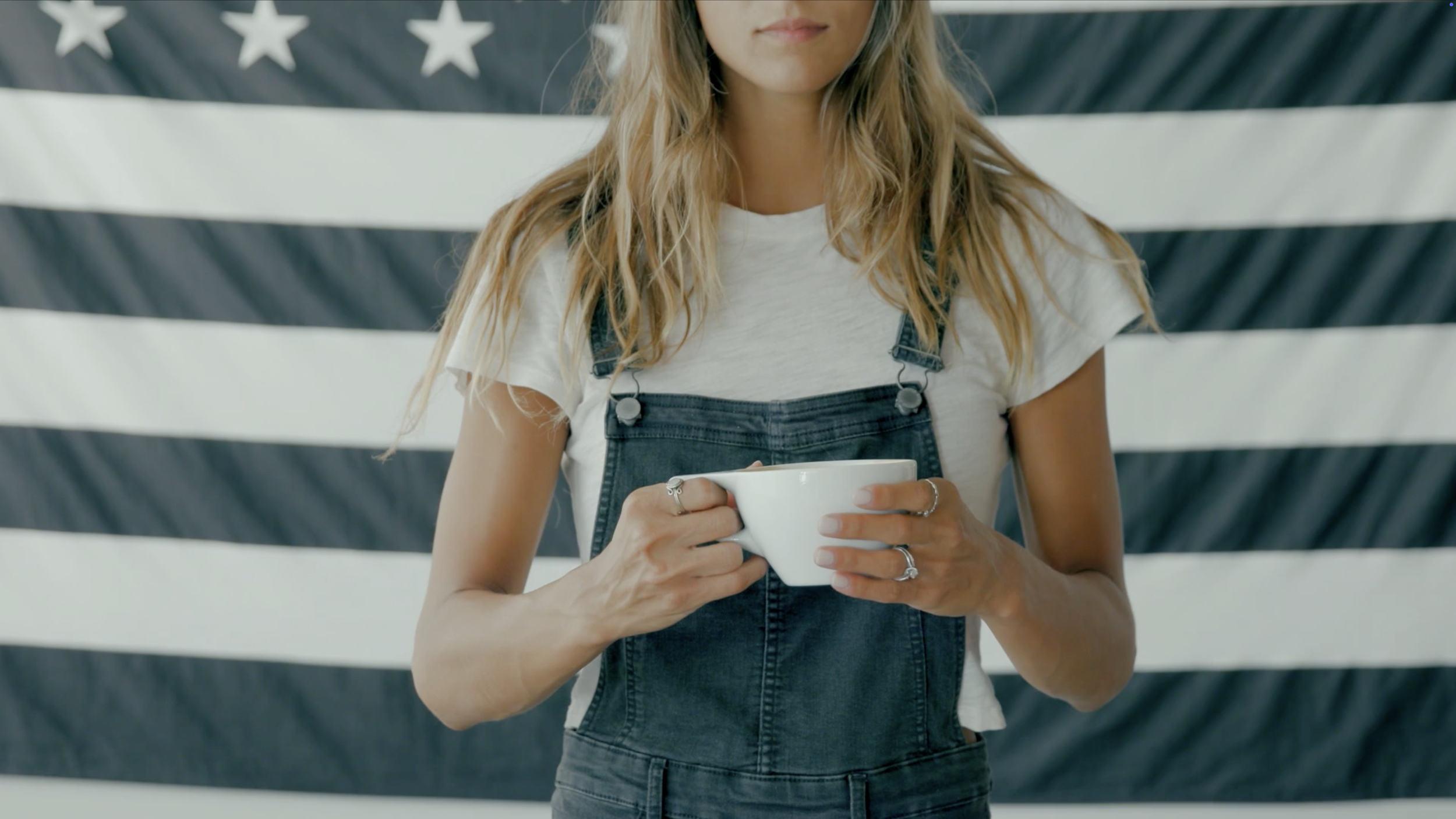 A woman with wavy blonde hair, wearing a white t-shirt and denim overalls, holding a white mug, standing in front of a black and white American flag wall.