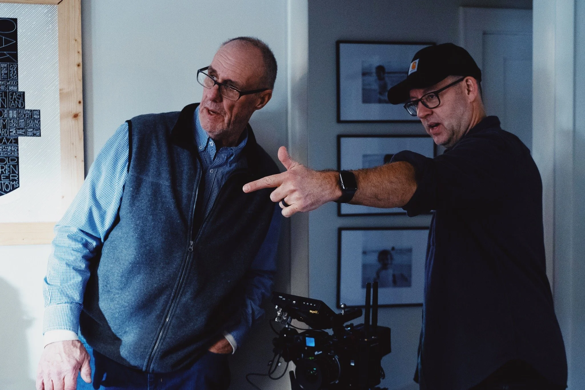 Two men with glasses and hats examining a camera or filming equipment in an indoor setting, with framed pictures on the wall in the background.