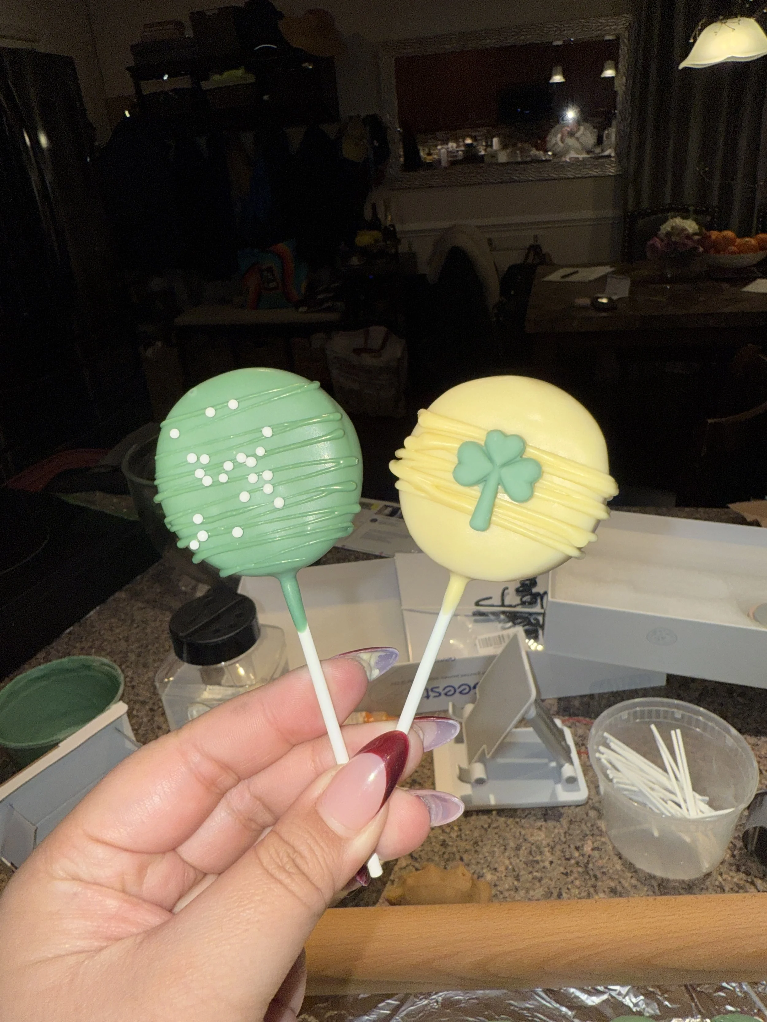 Two decorated cake pops, one green with white sprinkles and one yellow with a four-leaf clover decoration, being held in front of a kitchen counter.
