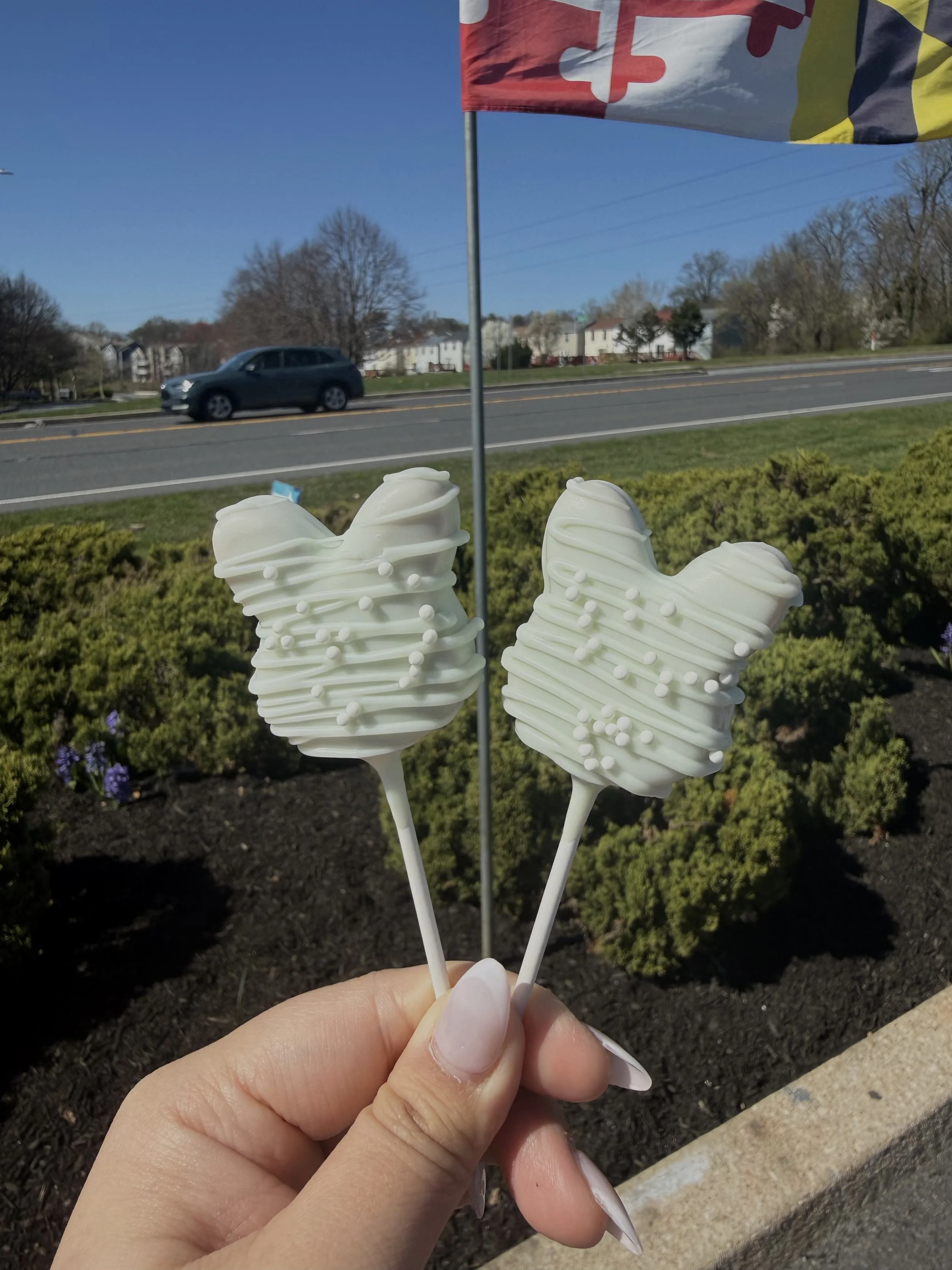 Two bunny-shaped marshmallow treats on sticks, decorated with white chocolate and sprinkles, held outdoors with a flag and a street in the background.