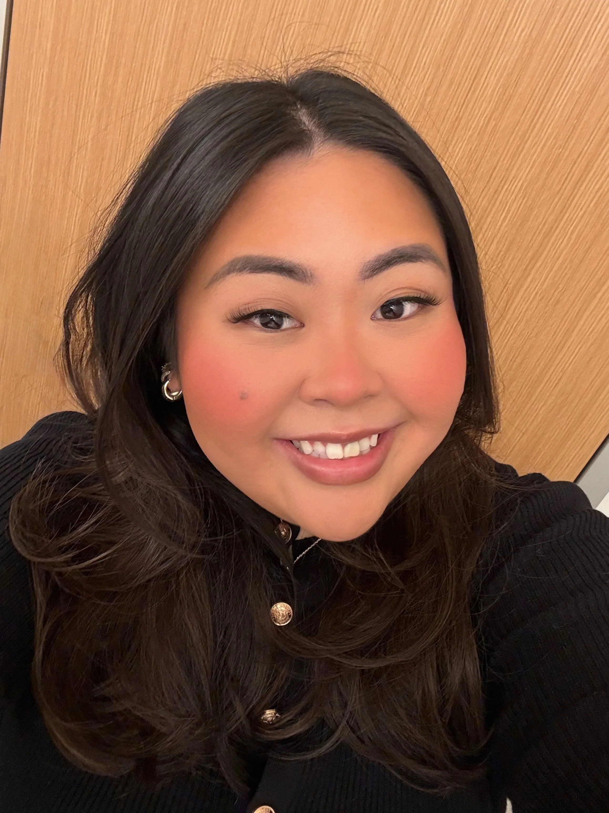 Close-up of a young woman with long dark hair, smiling, wearing a black top with gold buttons, and standing against a wooden background.