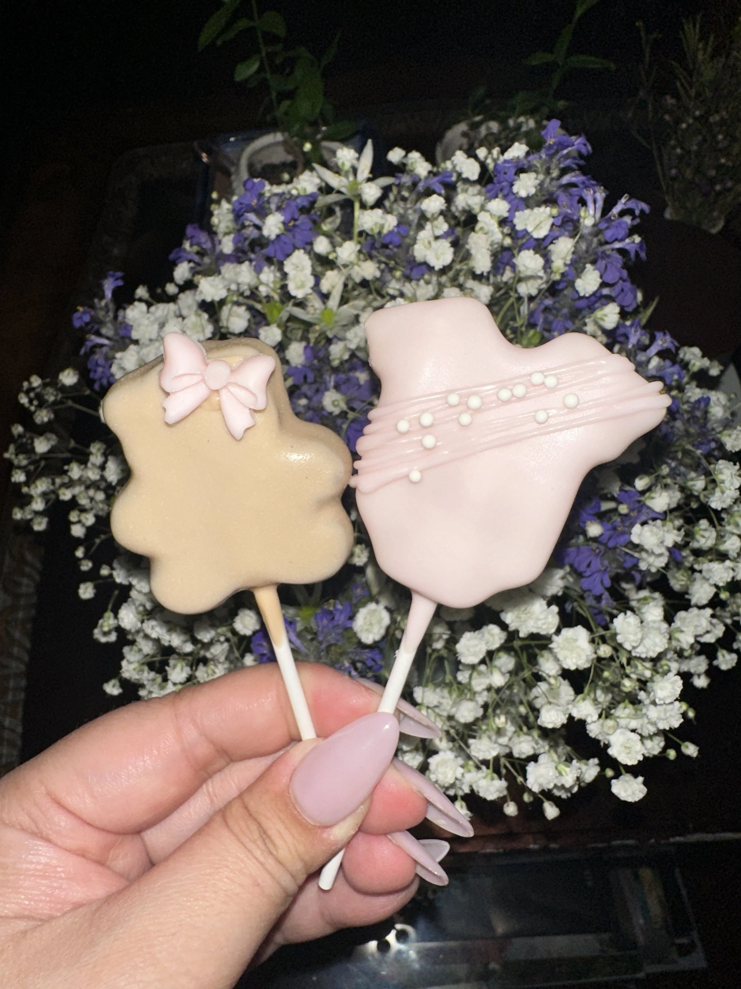 Two flower-shaped decorated lollipops, one beige with a pink bow and the other pink with white pearl accents, held in front of a bouquet of purple and white flowers.