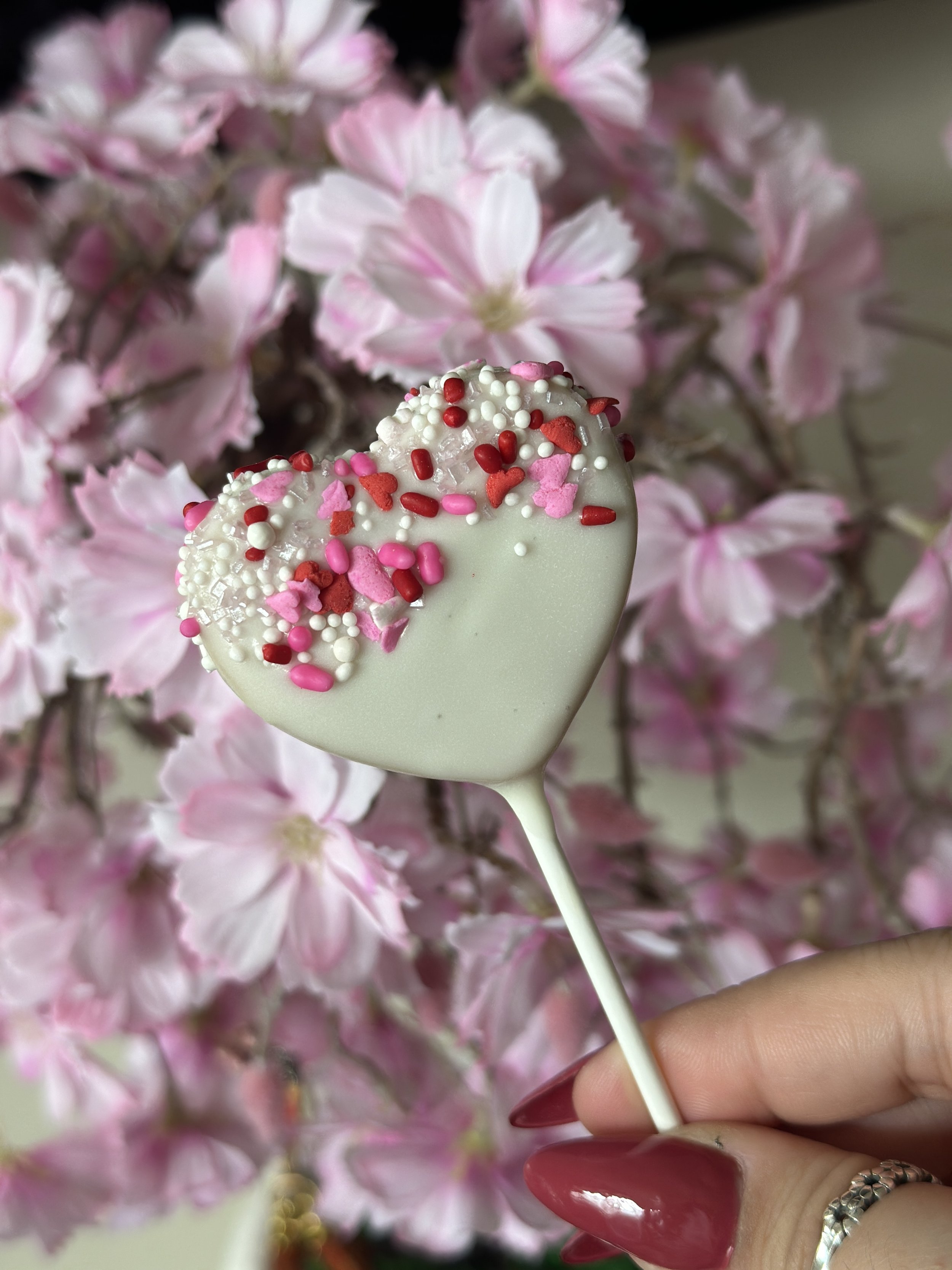 White heart-shaped lollipop with pink, red, and white sprinkles held by a hand with red painted nails, with pink flowers in the background.