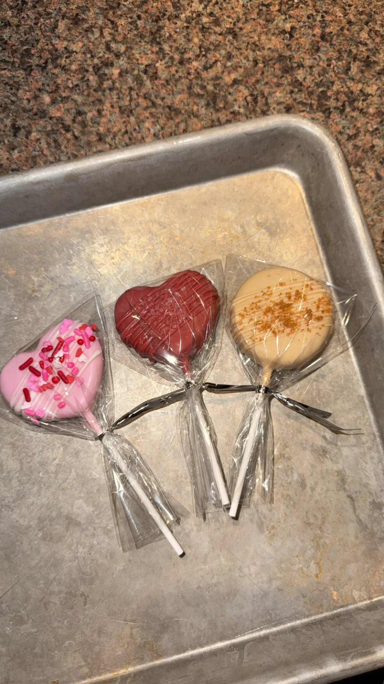 Three heart-shaped lollipops with pink, red, and white colors, decorated with sprinkles and caramel drizzle, wrapped in plastic, on a metal tray.