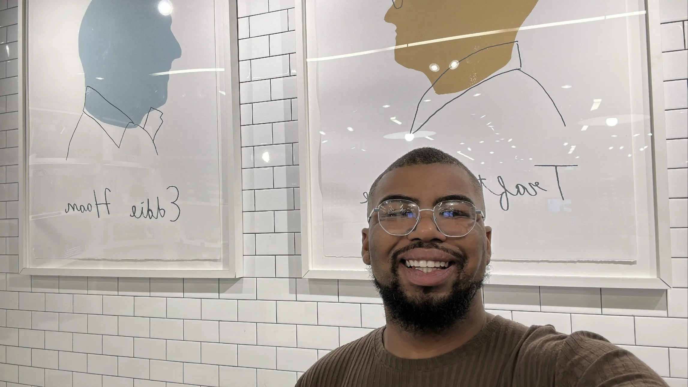 A smiling man with glasses taking a selfie in front of two framed illustrations of faces on a white tiled wall.