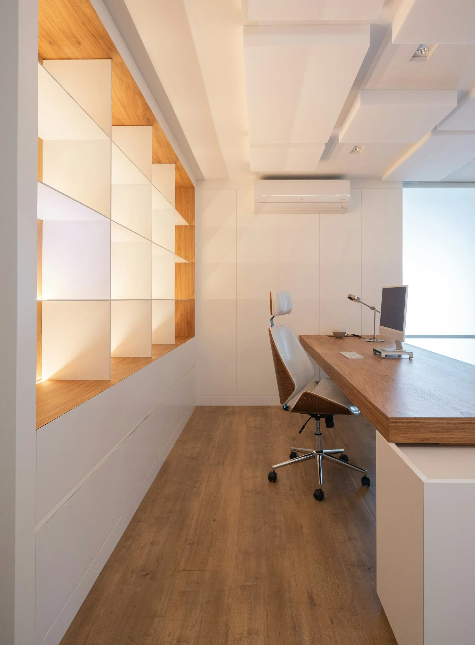 Minimalist office with a wooden desk, white ergonomic chair, computer monitor, desk lamp, and built-in wall shelves with white compartments and wood trim.