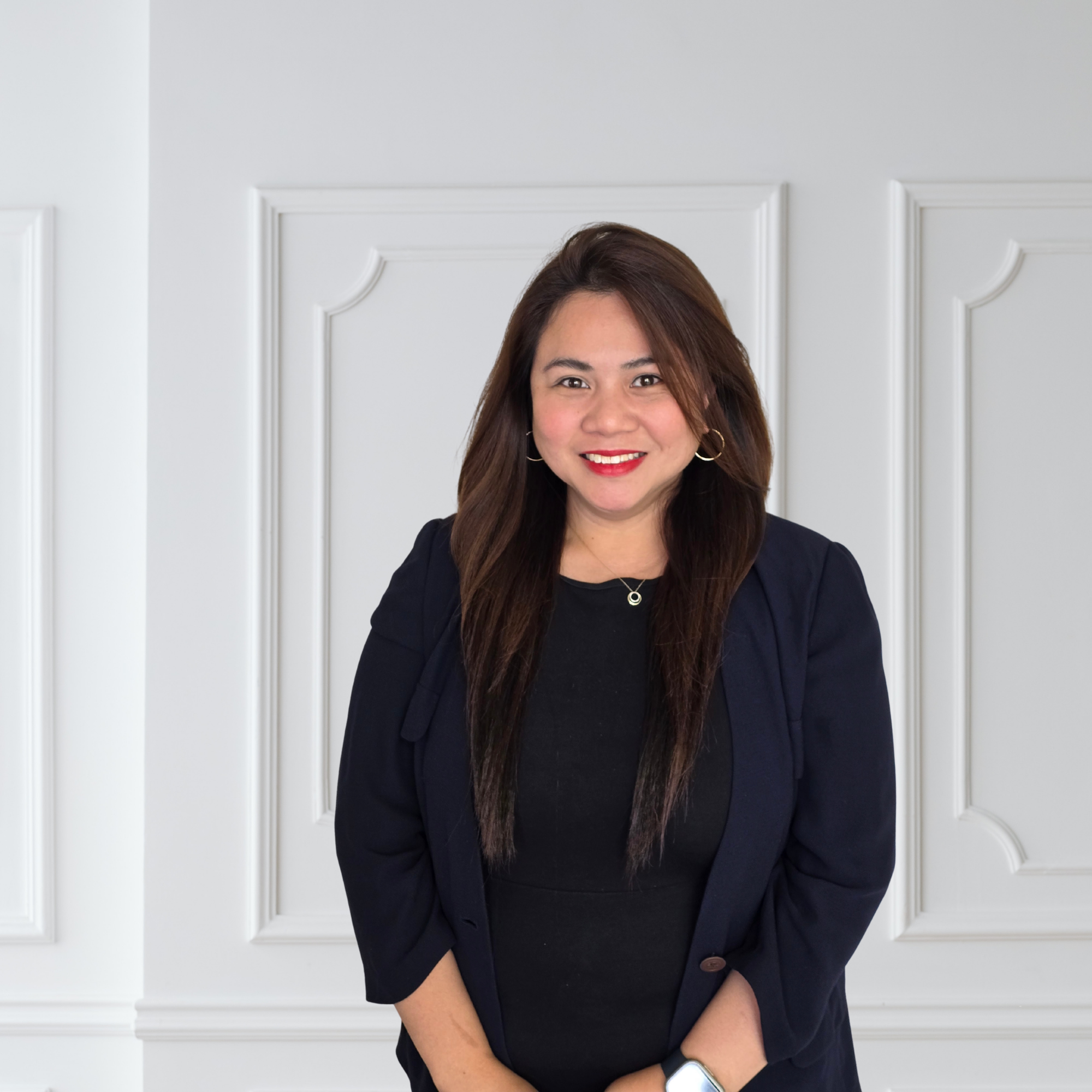 A woman with long brown hair wearing a black top and navy blazer, smiling, in front of a white paneled wall.