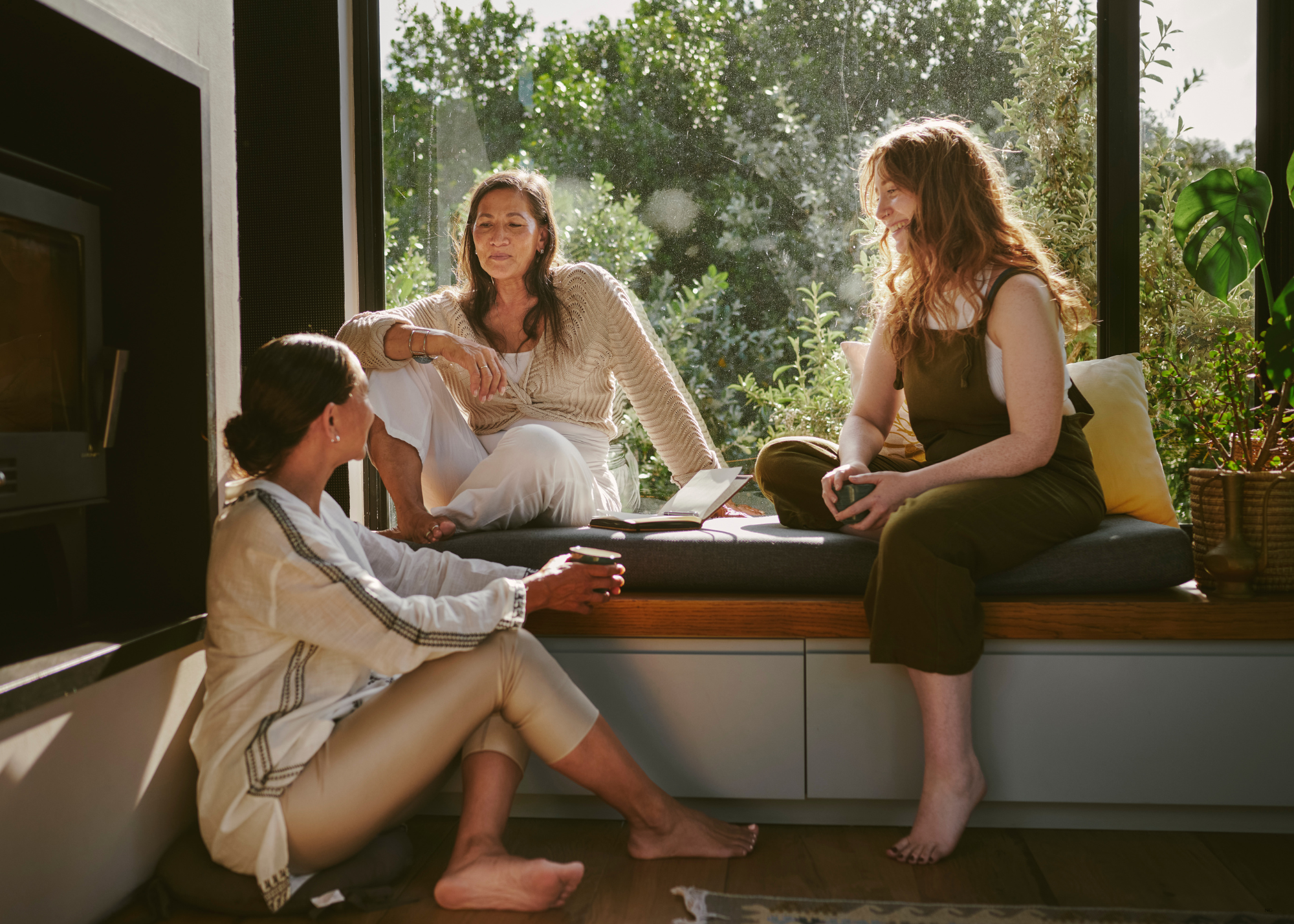 Four women sitting and relaxing in a bright, cozy room with a large window overlooking greenery, engaging in conversation and holding drinks.